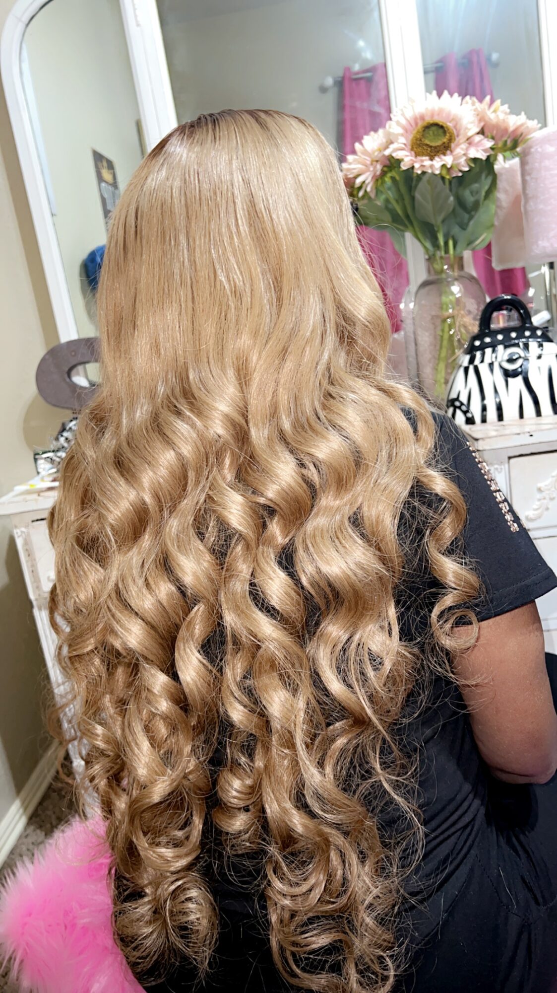Back of a person with long, blonde, curly hair in a room with a mirror and a vase of flowers