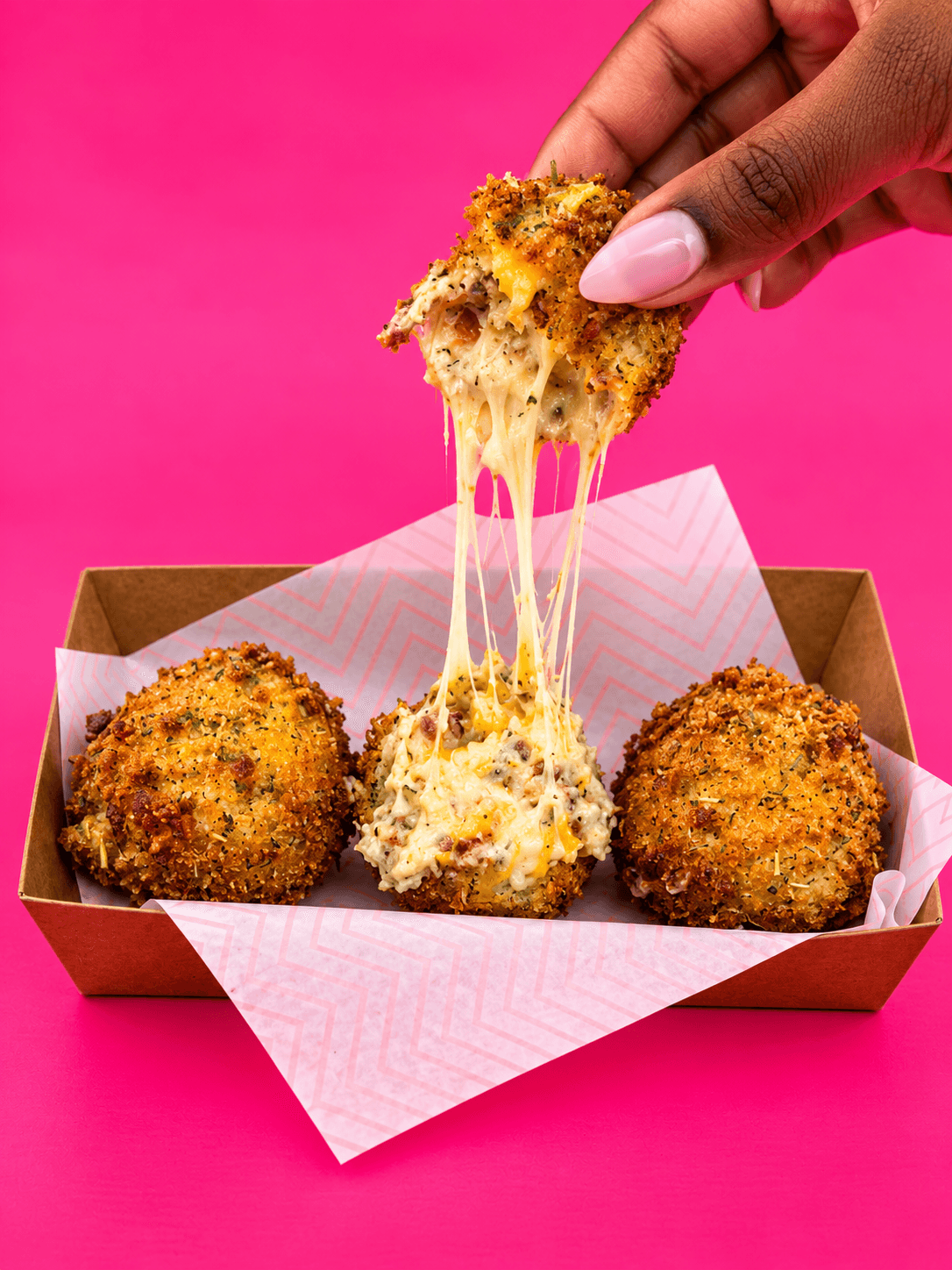 Hand pulling apart cheesy, breaded snack with melted cheese stretching, three similar snacks in a box on pink background.