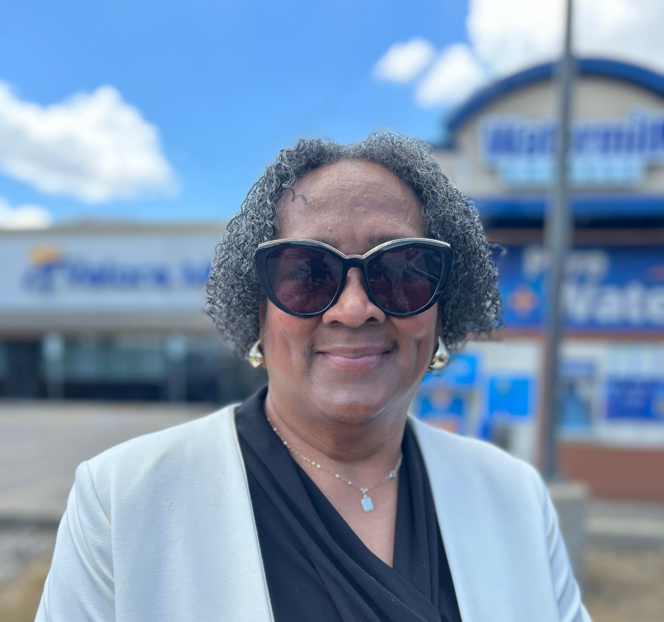 Woman with curly gray hair wearing sunglasses, a light blazer, and a black top, smiling outdoors.