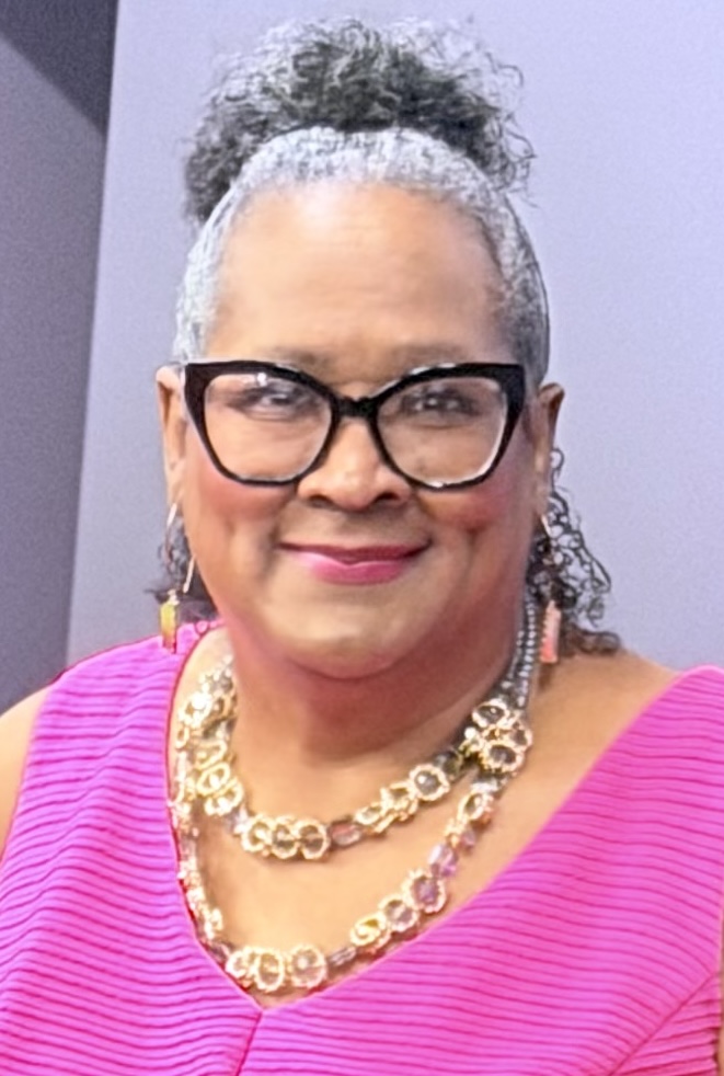 Woman with curly hair in a bun, wearing glasses, pink top, and gold jewelry, smiling.