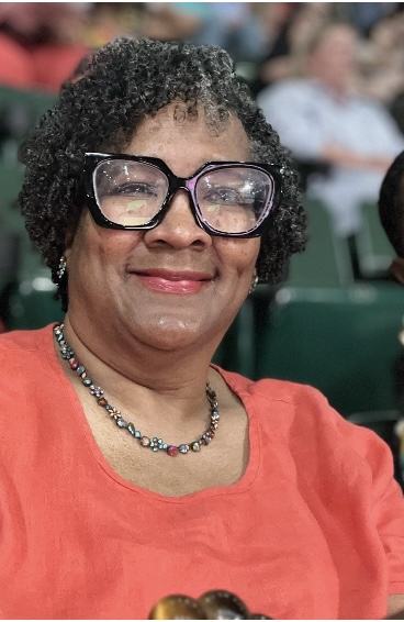 Woman with curly hair and glasses smiling, wearing a red top and a beaded necklace, seated in a crowd.