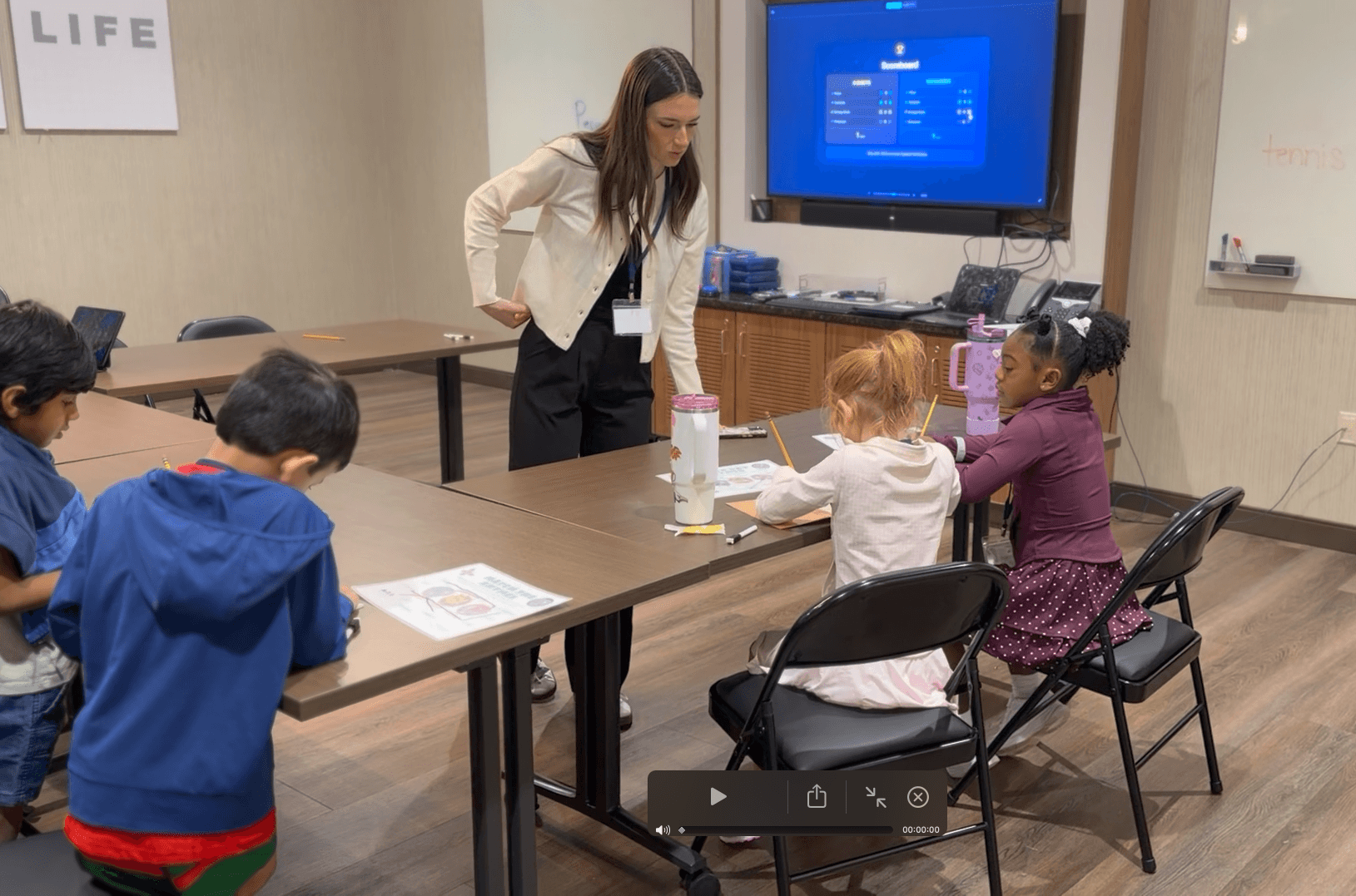 Teacher standing at a table with children seated, engaging in a classroom activity, with a screen and whiteboard in the background.