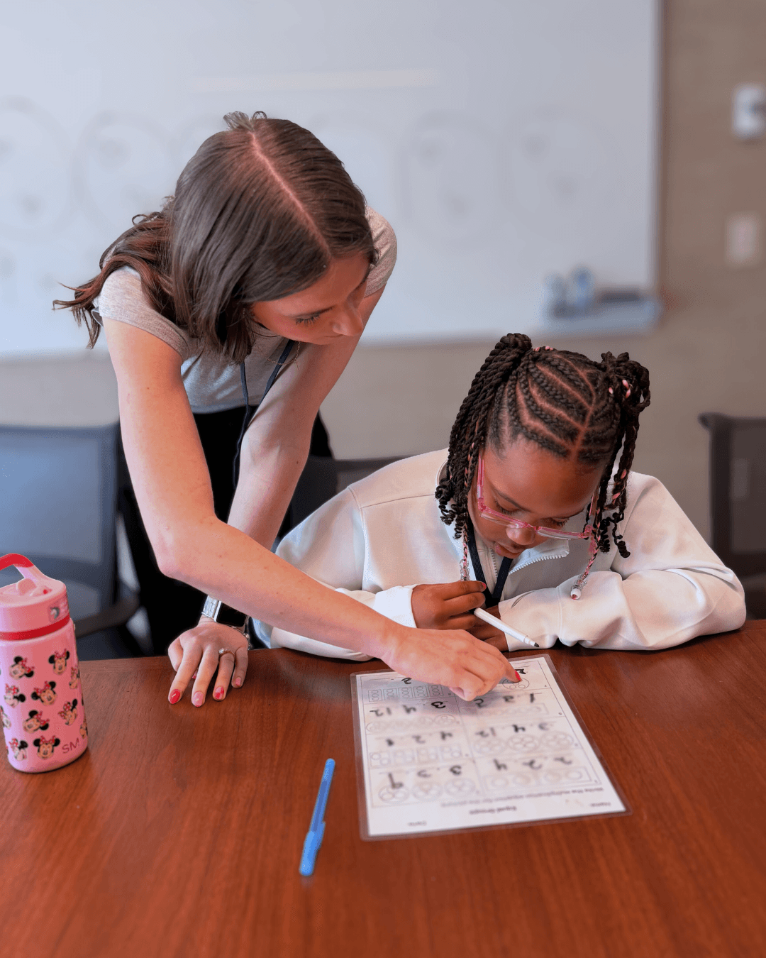Adult assisting child with worksheet at table, both focused on writing, classroom setting in background.