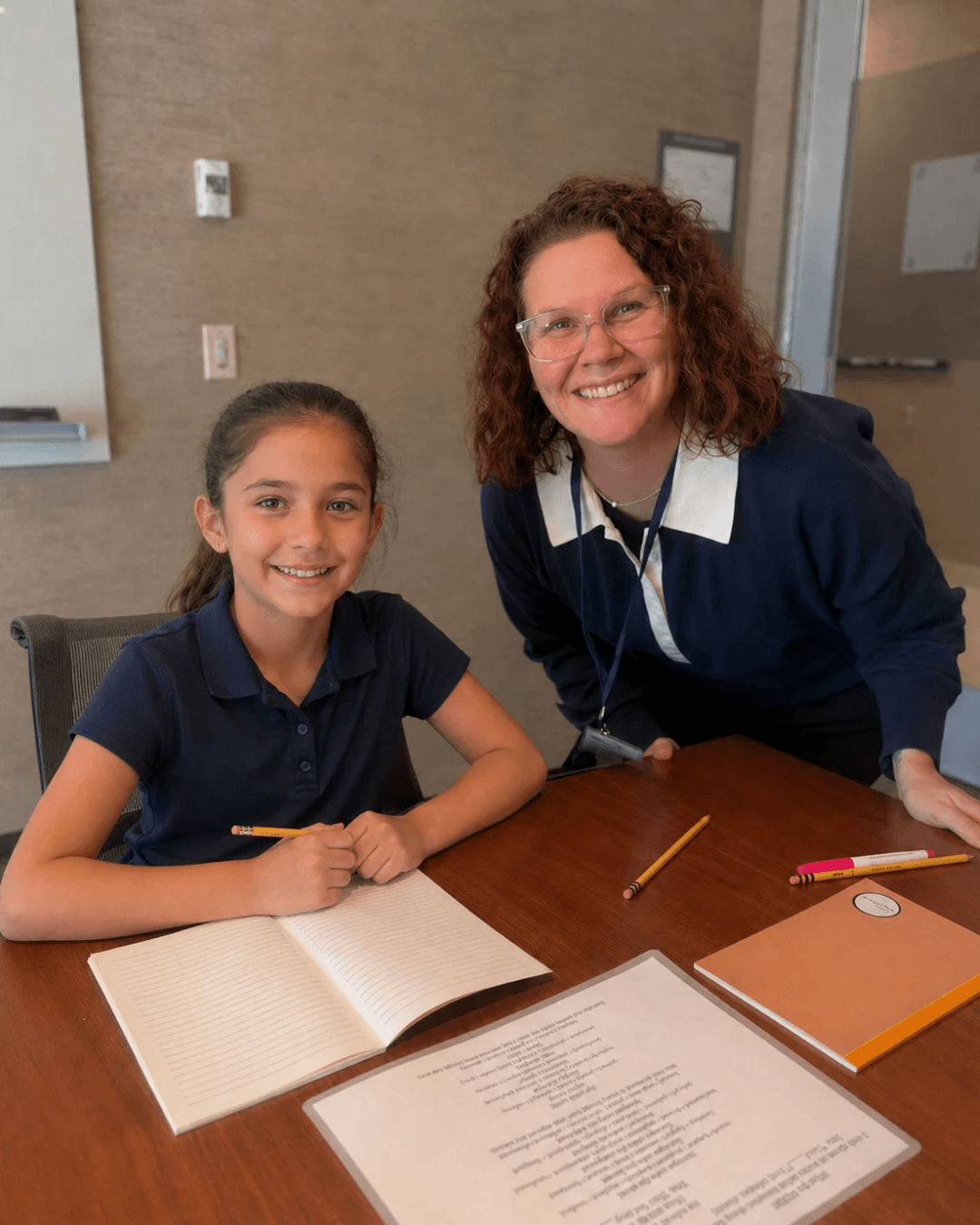 Two smiling females, one young girl and one adult woman, sit at a table with open notebooks and pencils, in a room with beige walls.