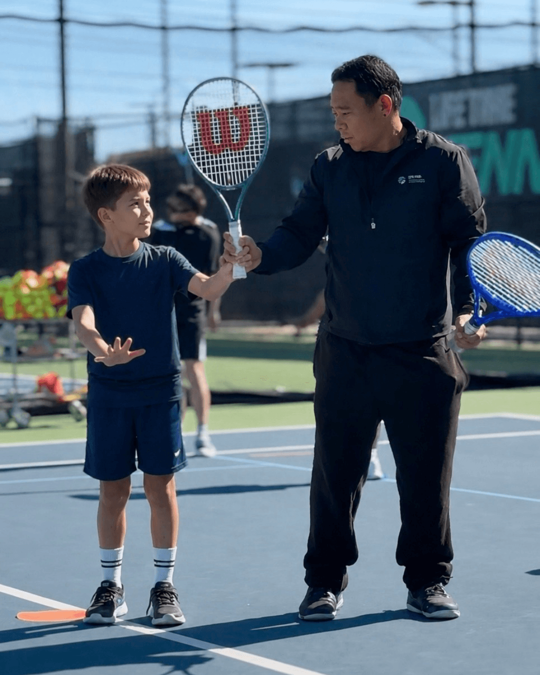 A young boy and an adult man shake hands on a tennis court, holding tennis rackets, with other players in the background.