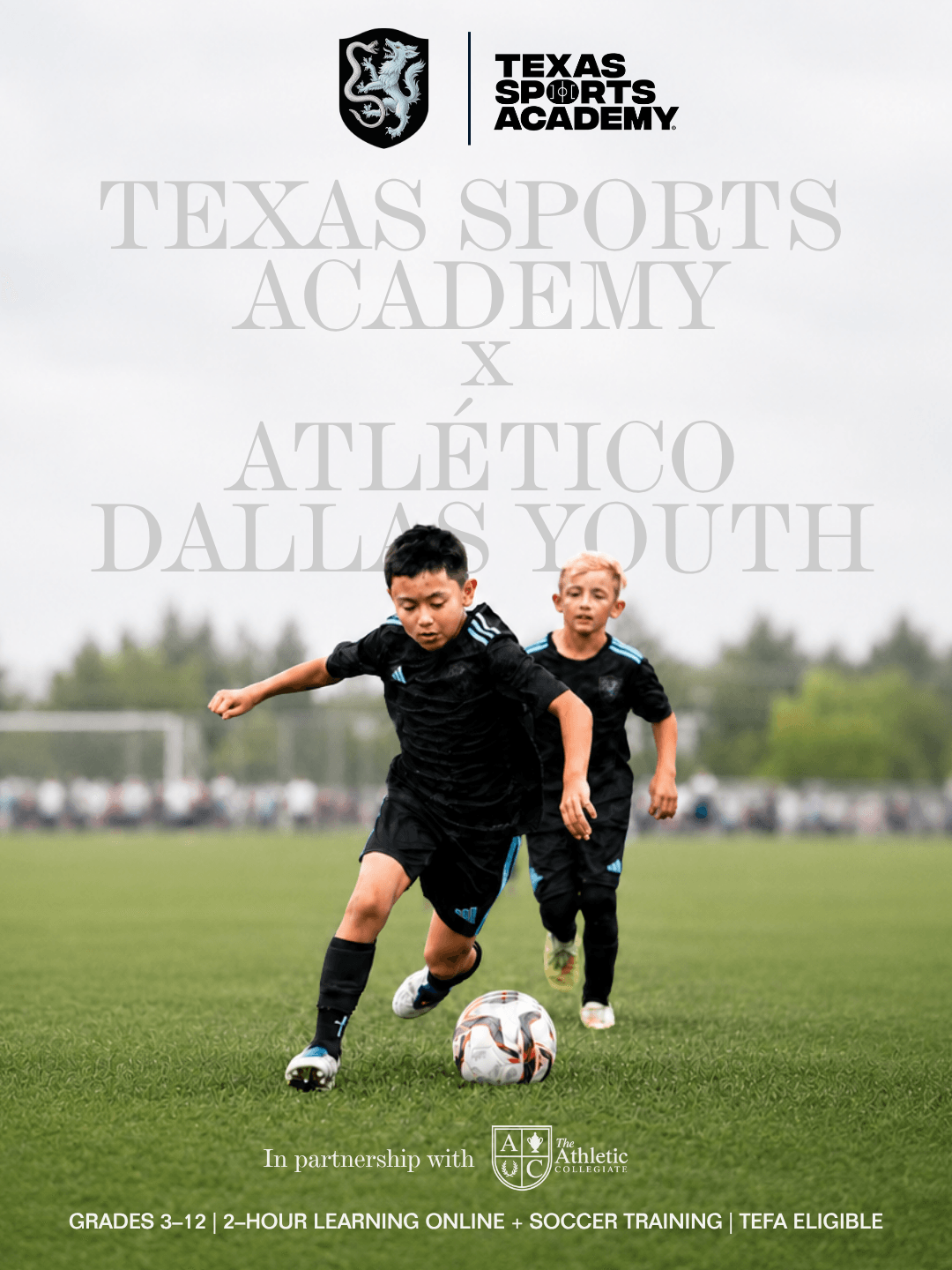 Two young boys playing soccer on a field, with a blurred background of trees and sky, wearing black sports uniforms.