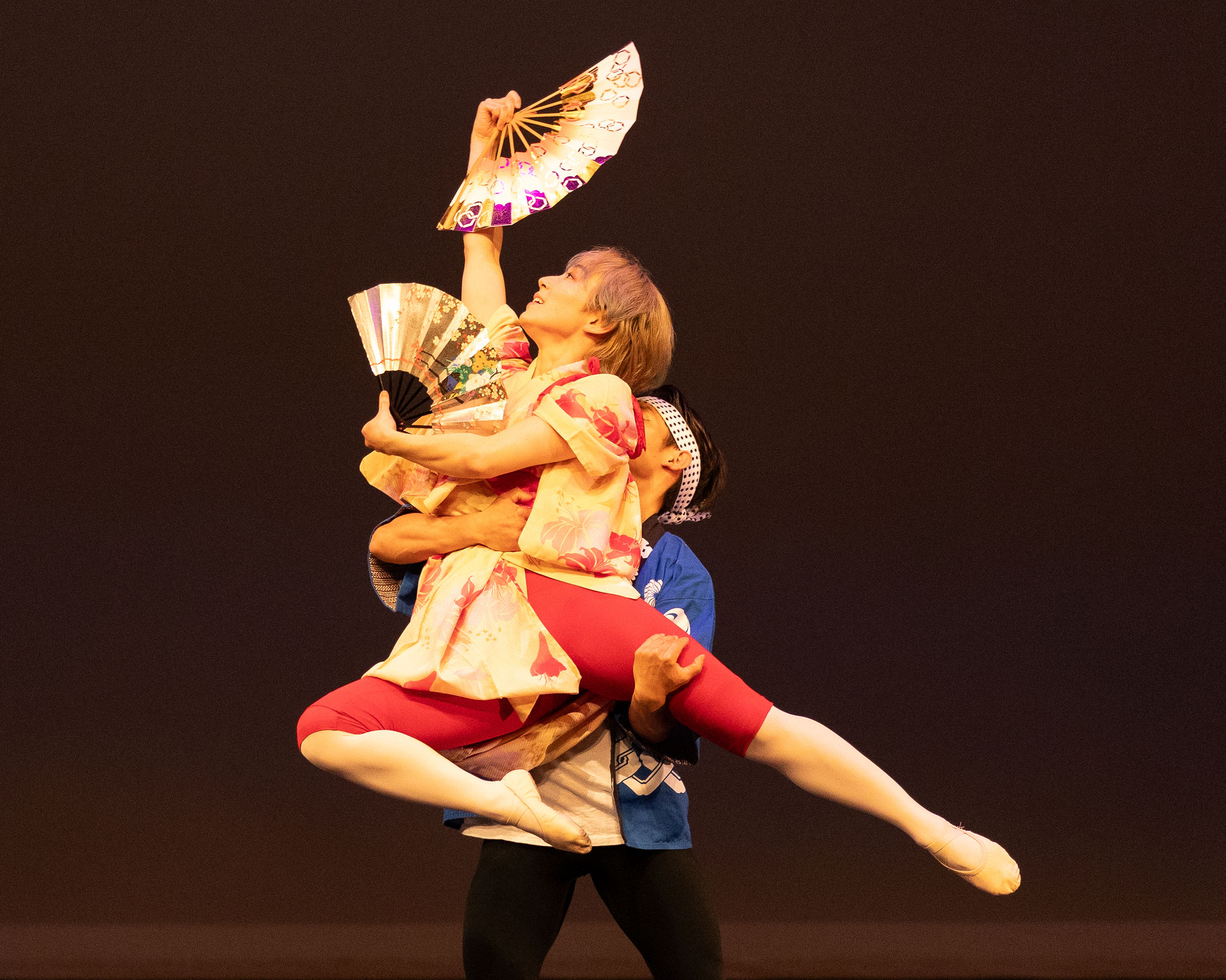 Two performers, one lifting the other, holding fans and a parasol, against a dark background.