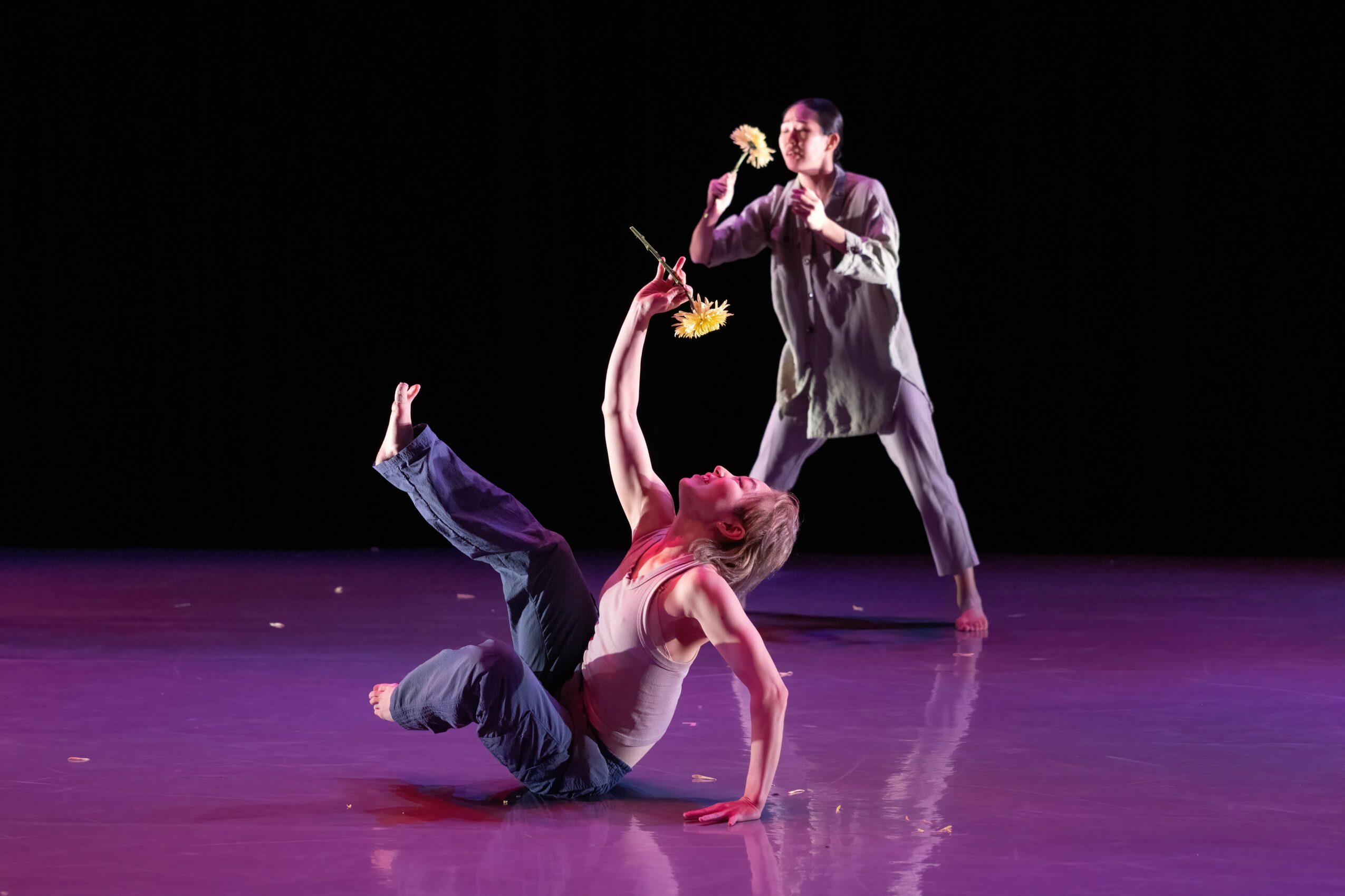 Two performers on stage, one lying on the floor and the other standing, both engaged in a dance or performance.