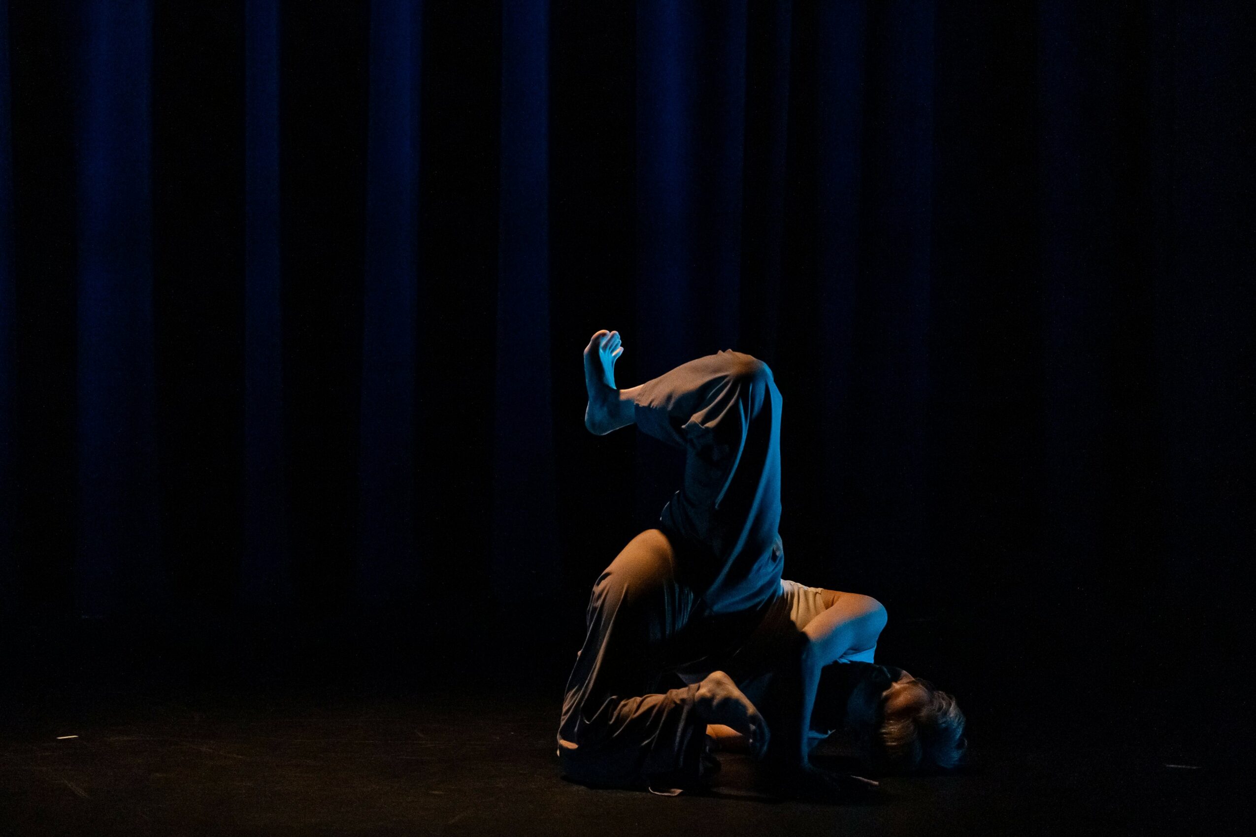 Person performing a dance move on stage with dark curtains in the background.
