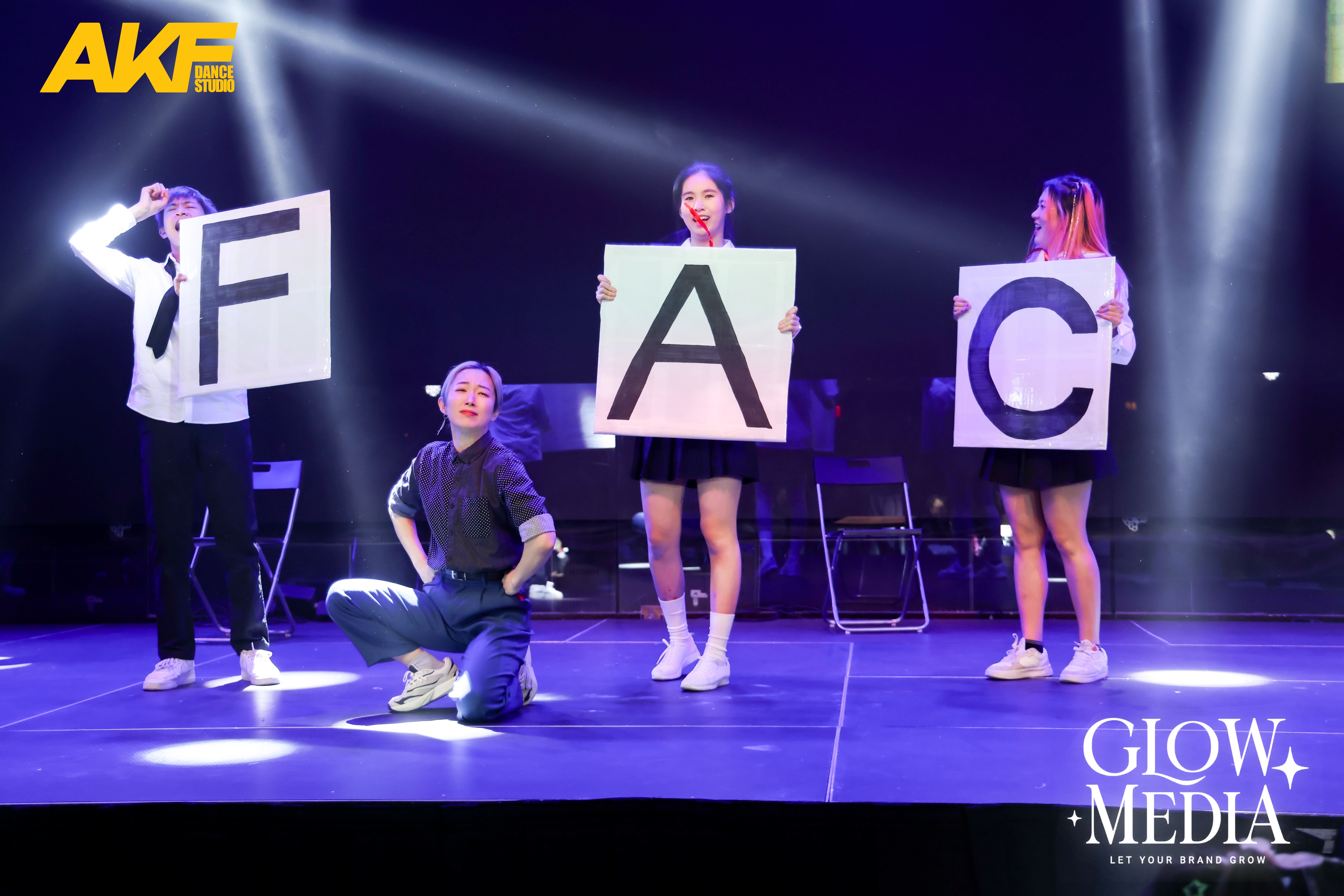 Five people on stage holding large letters spelling 'FAC', with stage lights and a dark background.