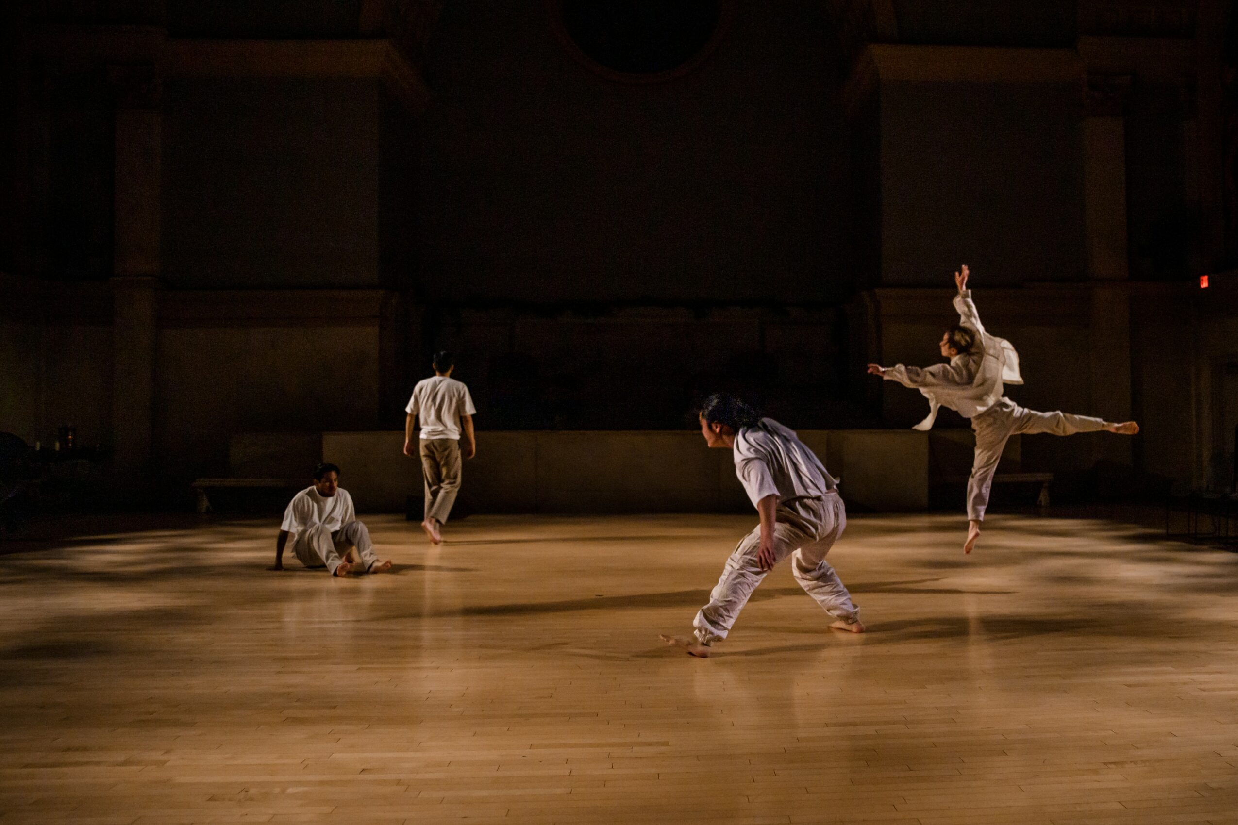 Four dancers perform on a wooden stage in a dark theater, with one dancer mid-air in a leap.