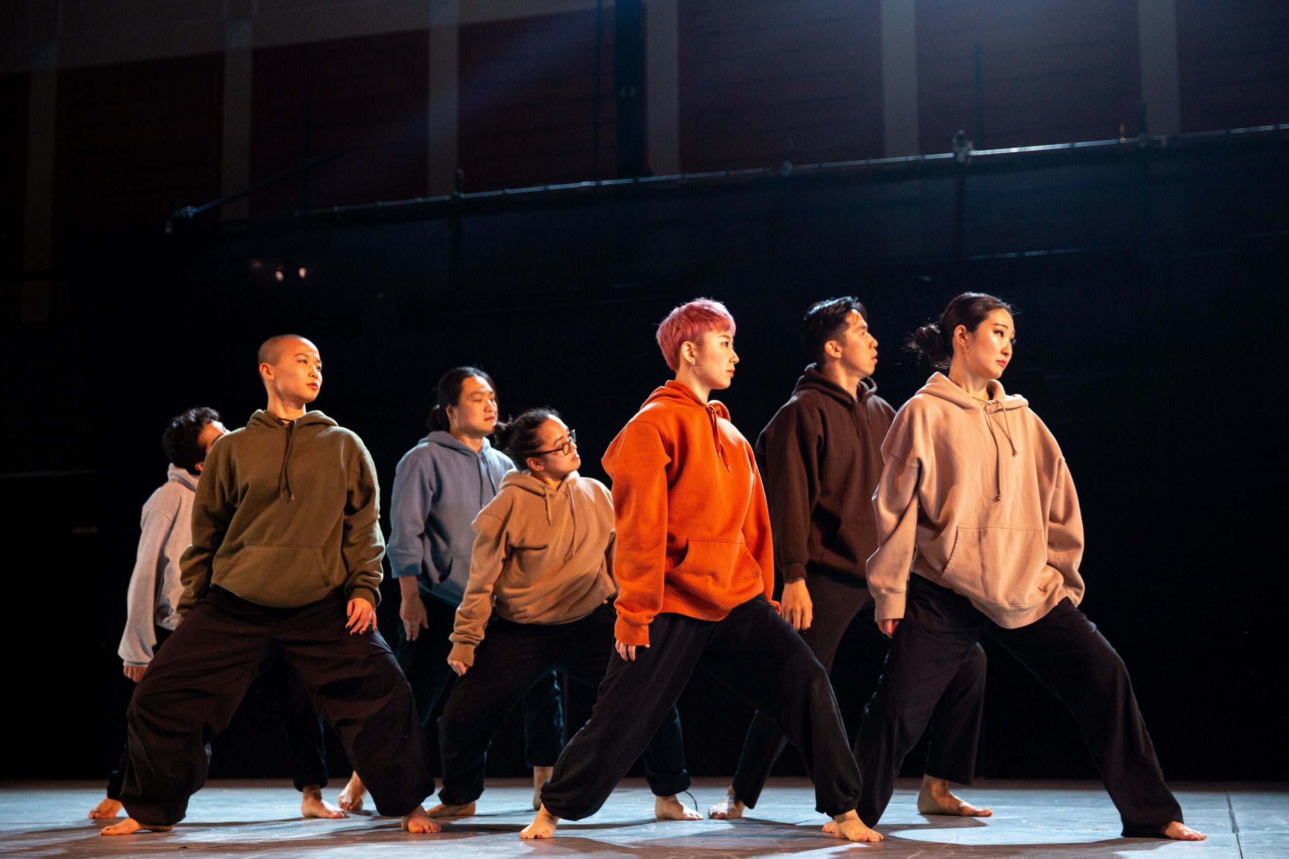 Group of people performing a dance or exercise on stage, facing different directions, with a dark background.