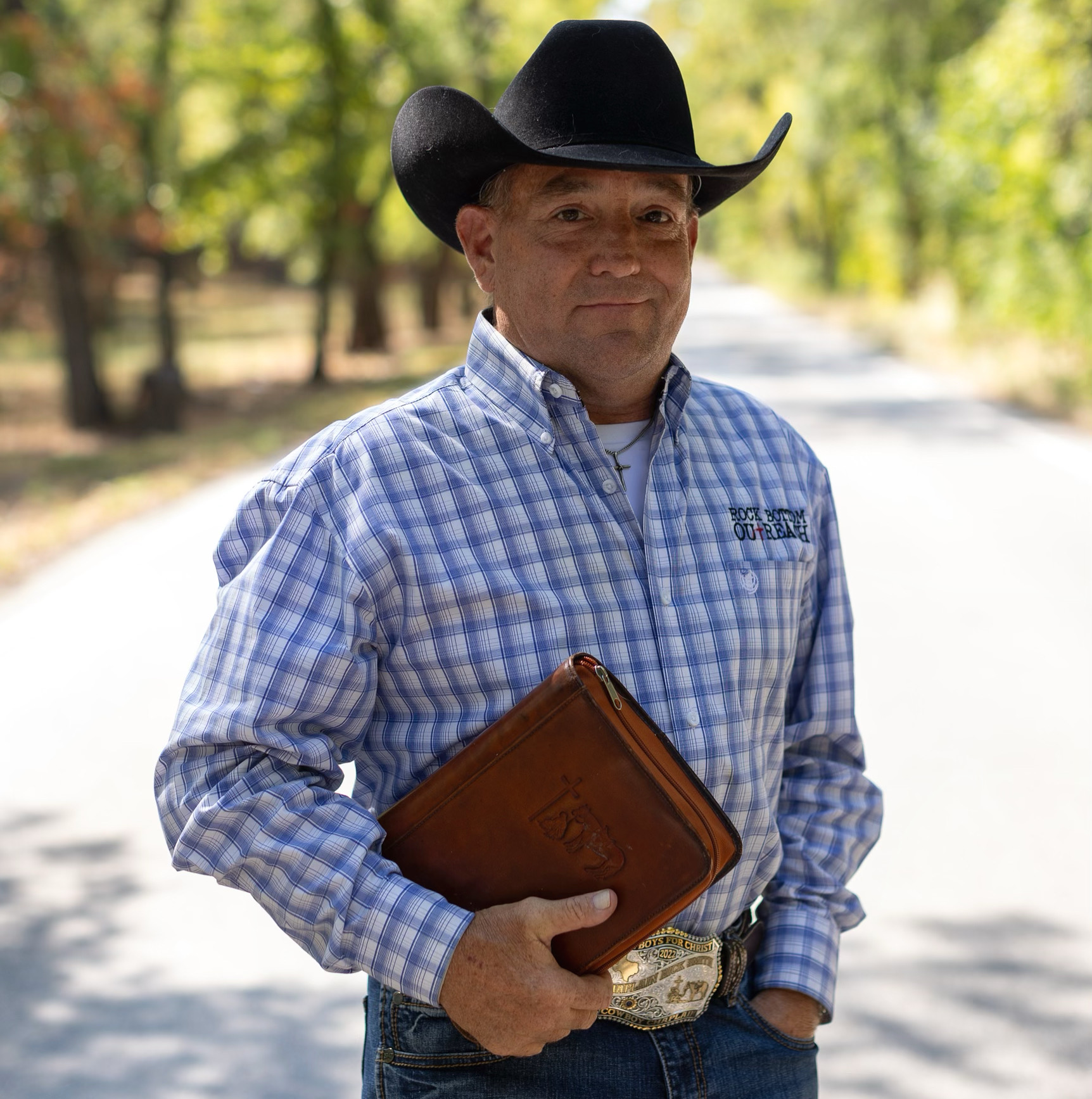 Man wearing a cowboy hat and checkered shirt holding a brown wallet outdoors on a sunny day.