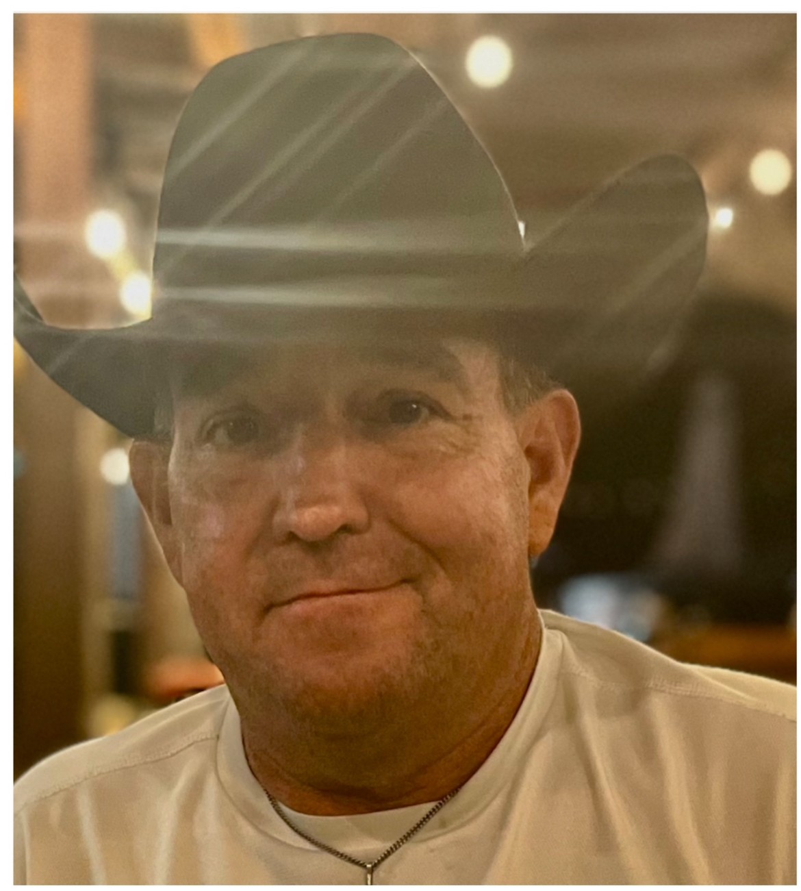 Man wearing a cowboy hat smiling, indoor setting with warm lighting, blurred background, casual attire.