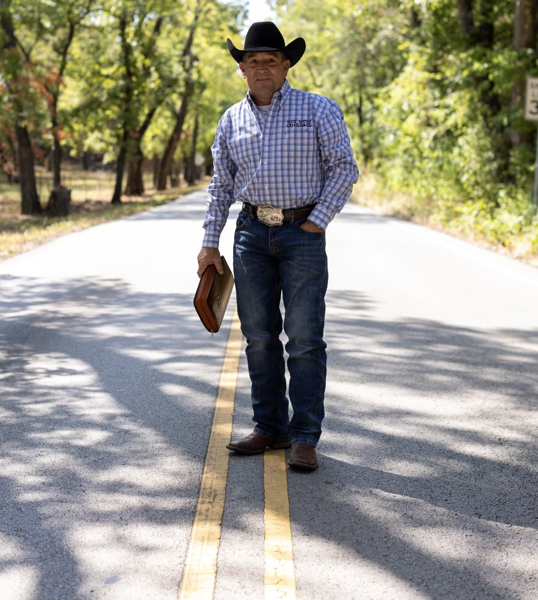 Man in cowboy hat and checkered shirt standing on a rural road holding a hat, trees in background.