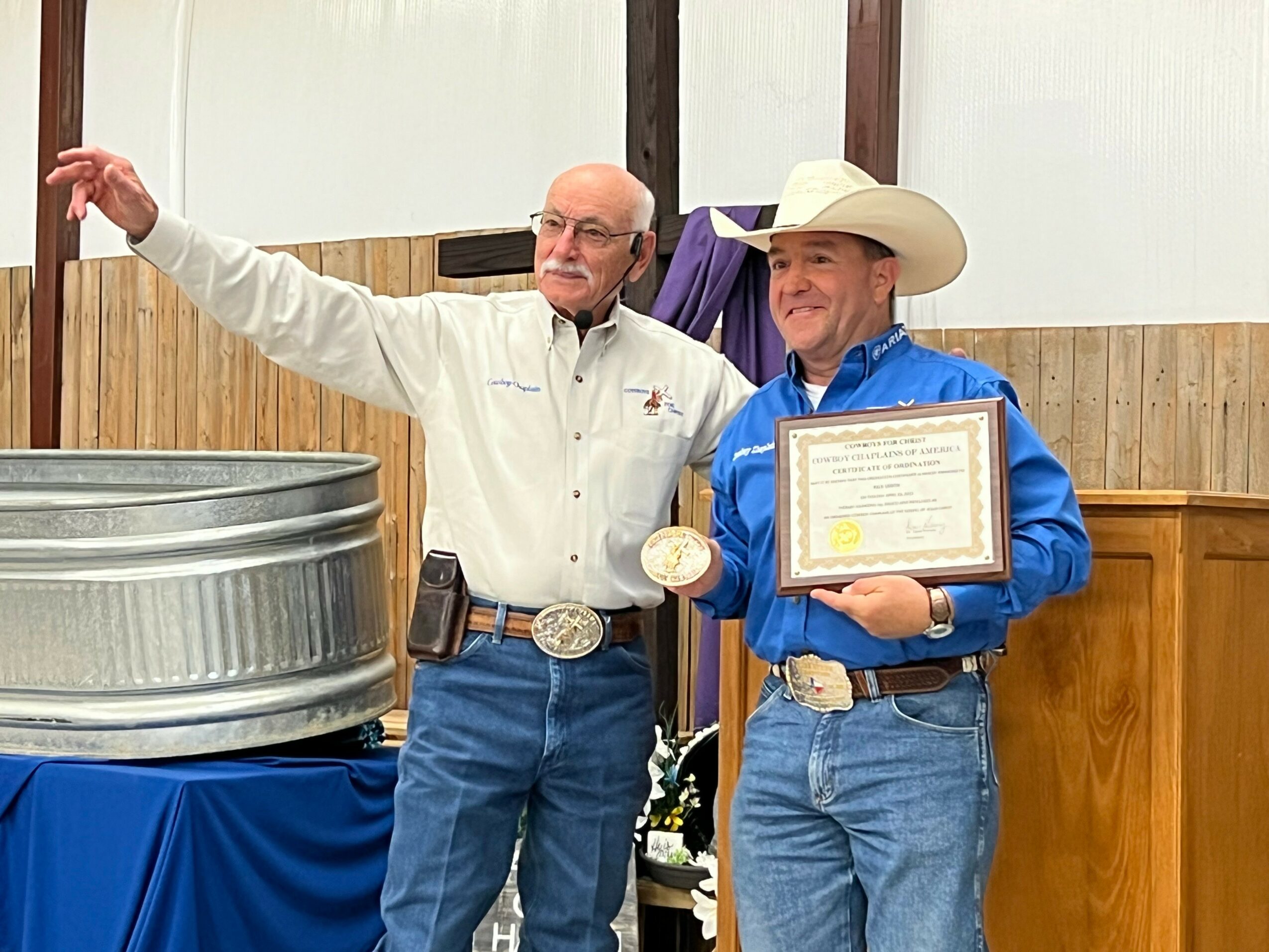 Two men stand indoors, one holding a framed certificate and a medal, the other gesturing with his arm. Both wear cowboy hats.