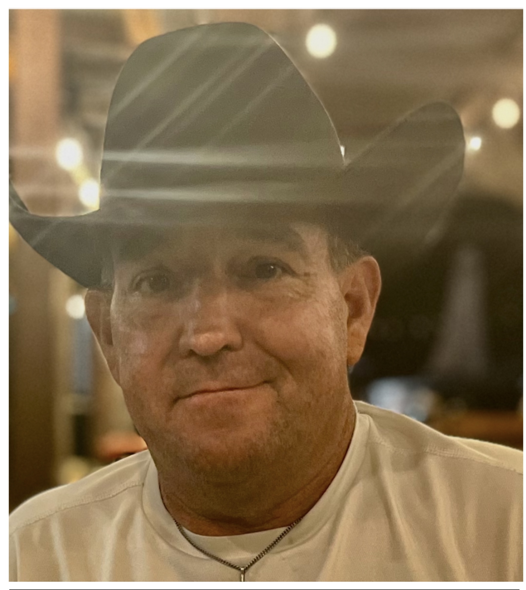 Man wearing a cowboy hat and white shirt, smiling, in a warmly lit indoor setting.