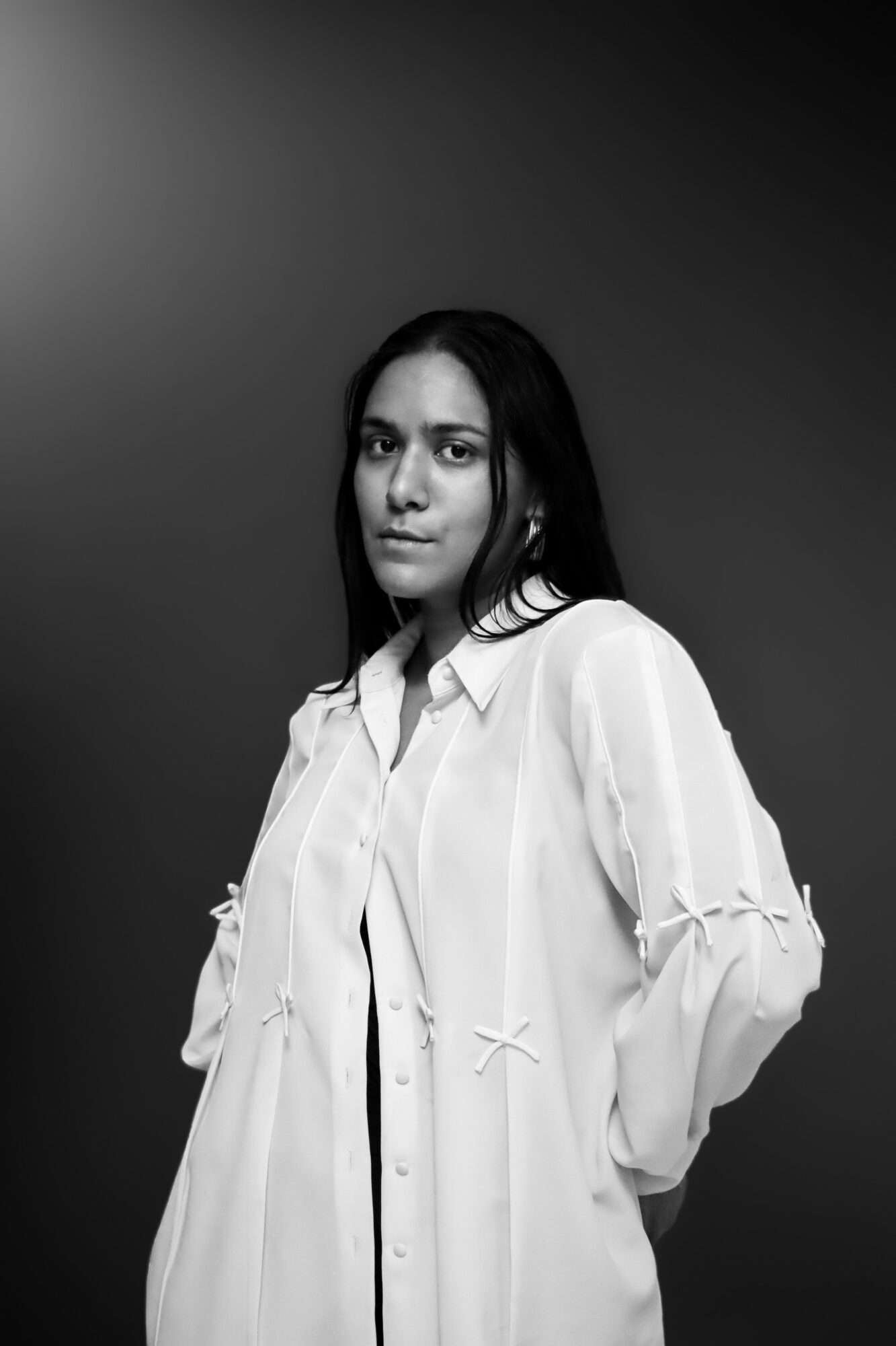 Woman with long dark hair wearing a light-colored shirt, standing against a dark gradient background.