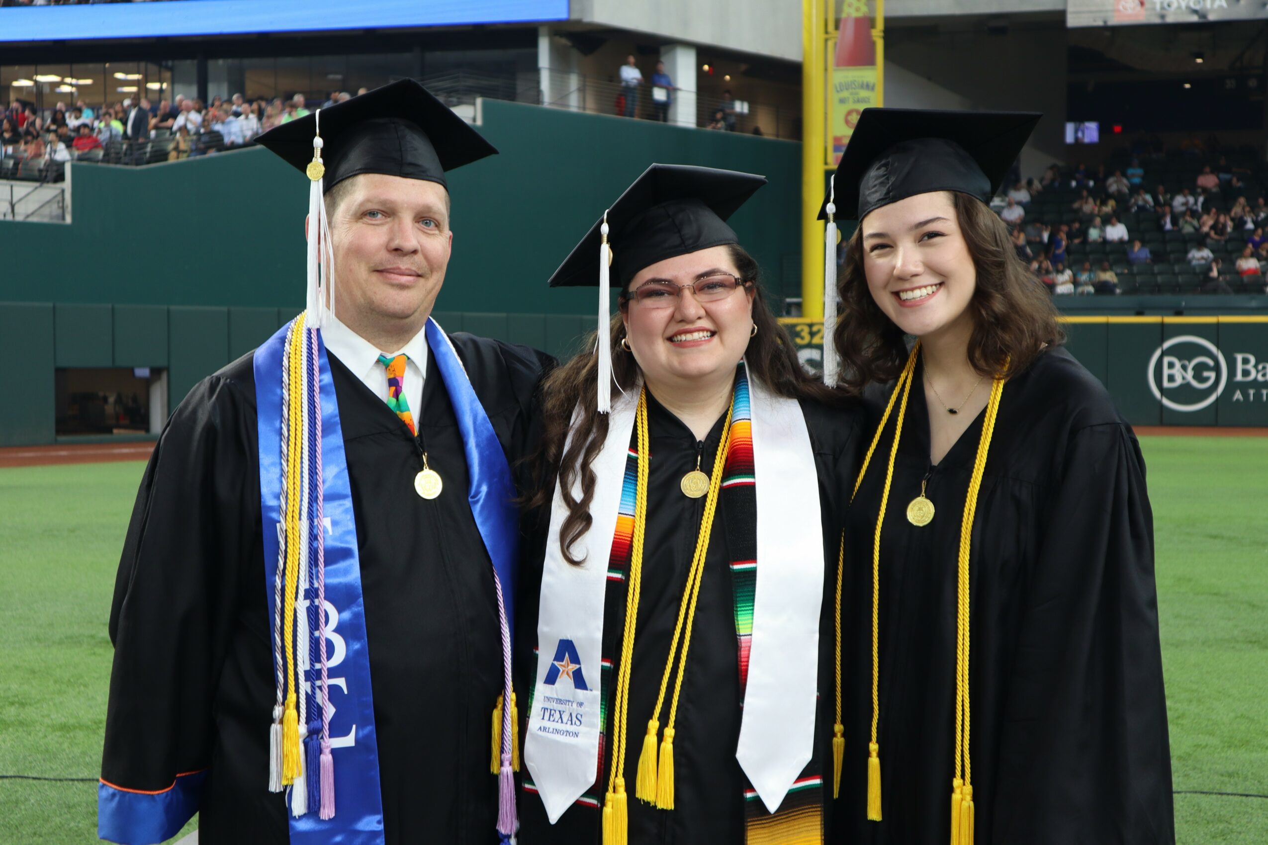 Three graduates in caps and gowns standing on a field, smiling, with a crowd in the background.