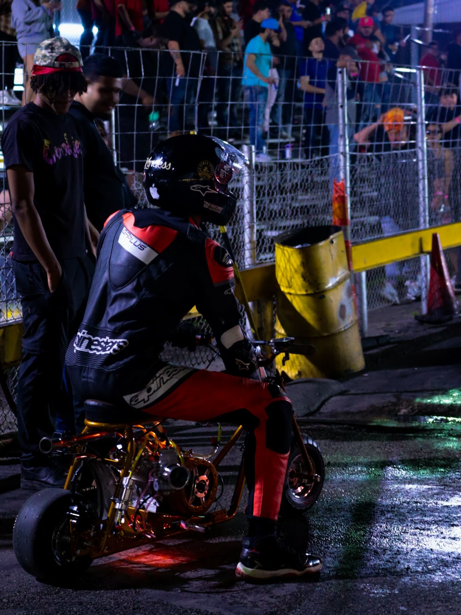 Person in racing gear sitting on a small motorcycle at night, with a crowd and barriers in the background.