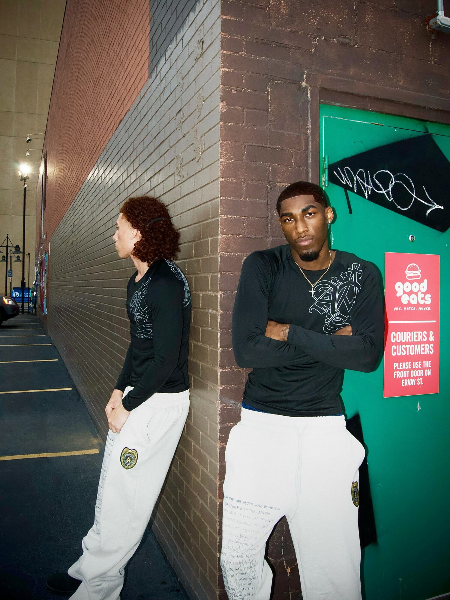 Two people stand against a brick wall and a teal door, one facing sideways, the other with arms crossed, in an urban setting.
