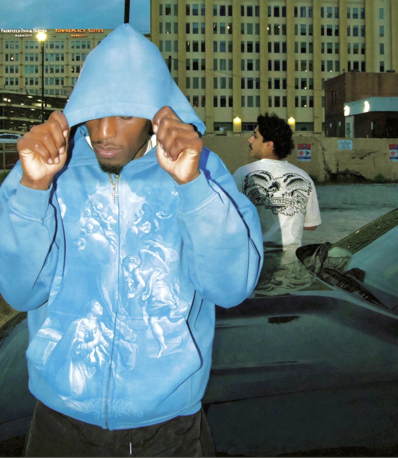Person in blue hoodie adjusting hood, cityscape background with buildings and a parked car.