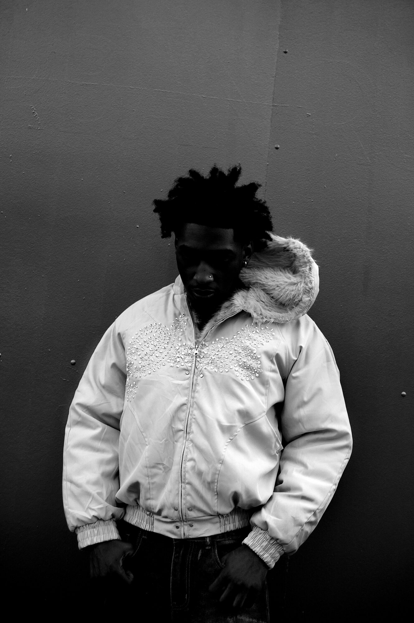 Person with curly hair wearing a light-colored jacket with a fur-lined hood, standing against a plain wall.