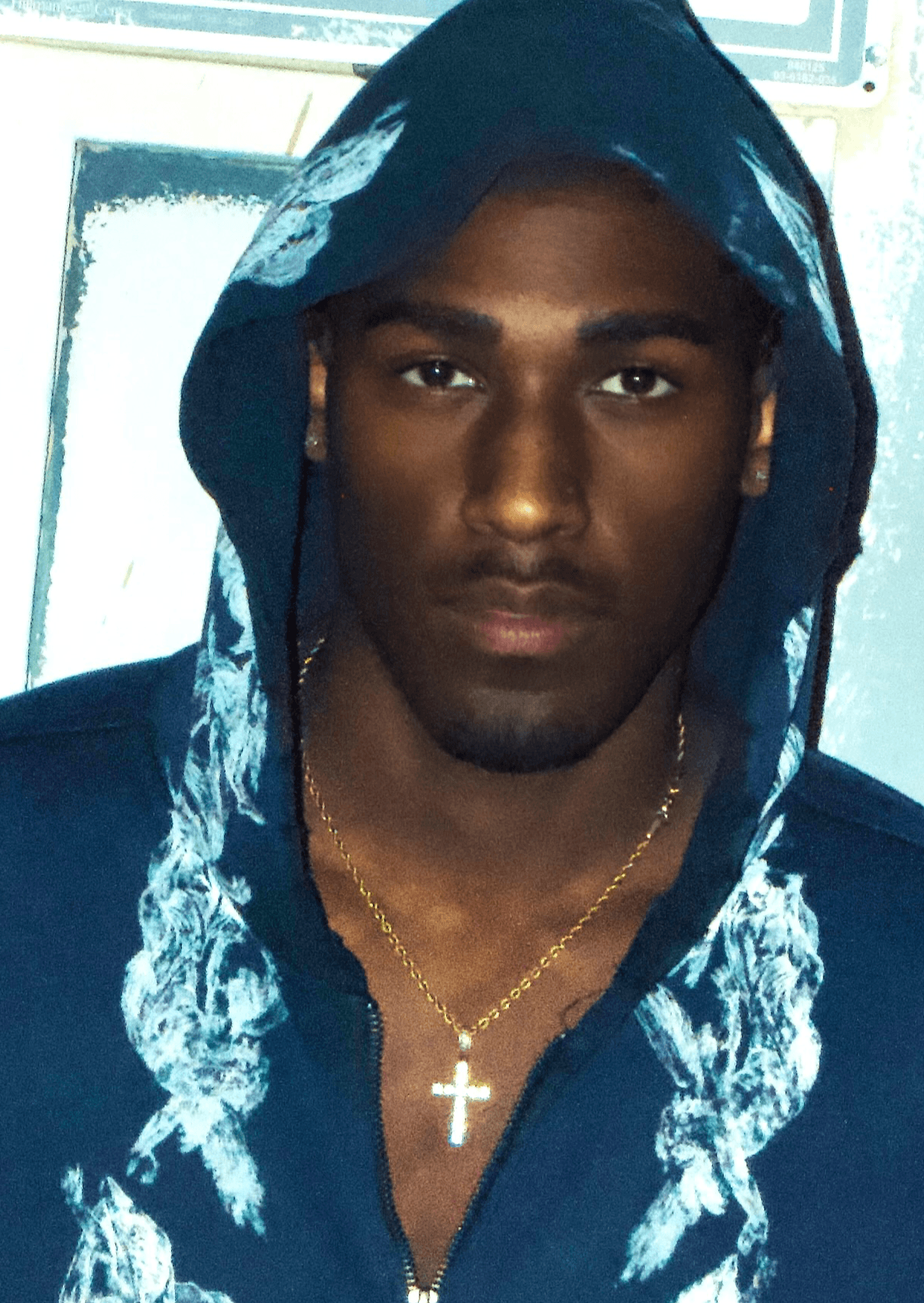 Young man with a hood, wearing a gold cross necklace, looking directly at the camera.