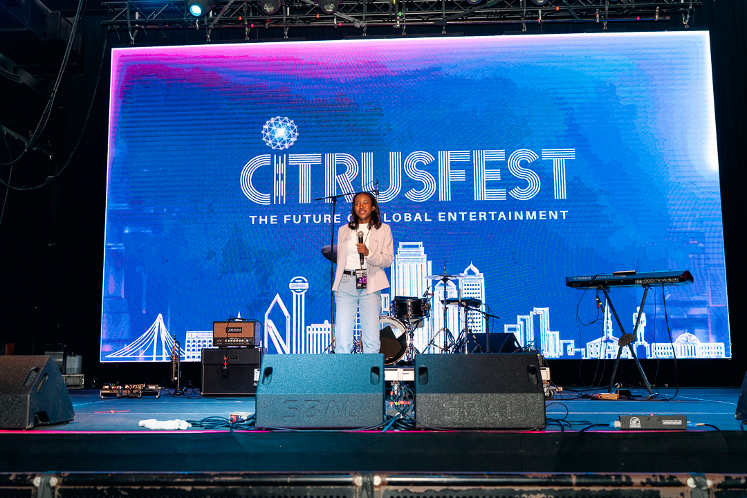 A woman stands on stage with a large digital screen behind her displaying 'CITRUSFEST' and cityscape graphics.