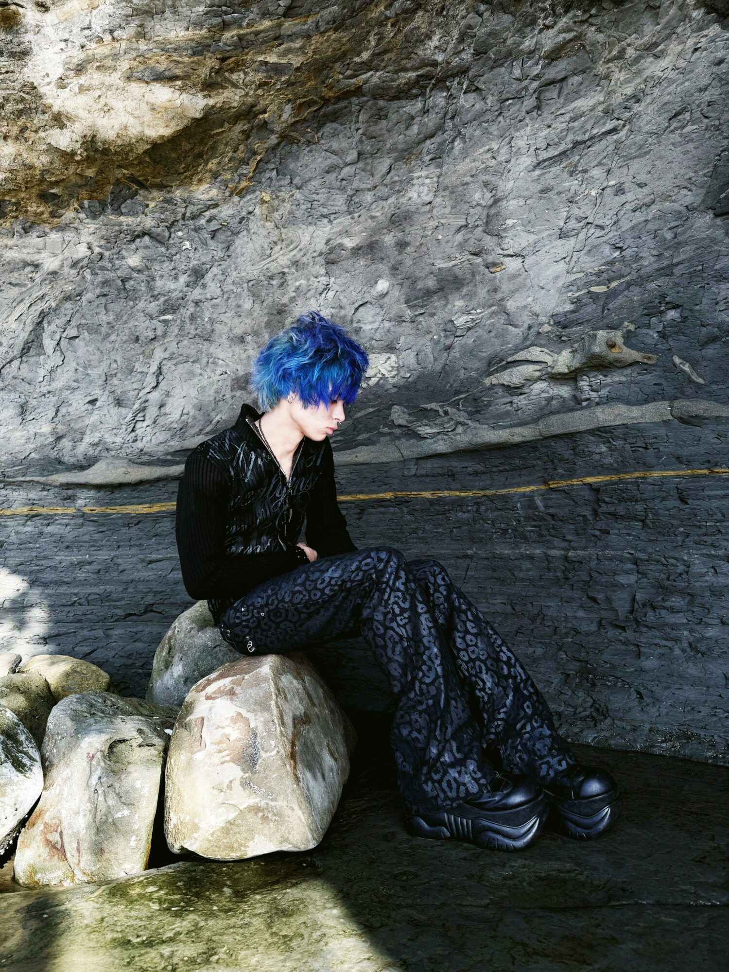 Person with blue hair sitting on rocks inside a cave, looking down, wearing black clothing and patterned pants.