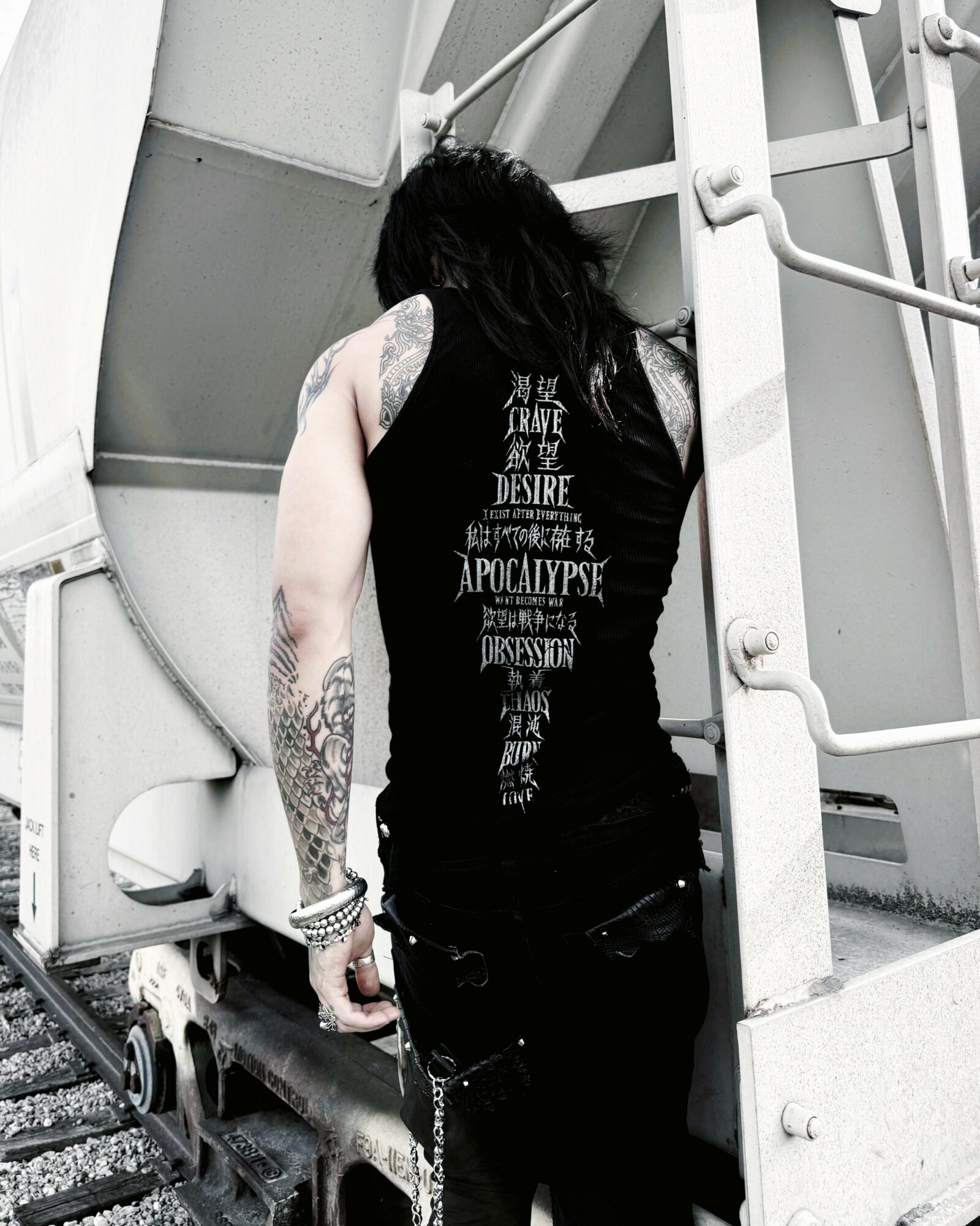 Person with long dark hair and tattoos on arm, wearing a black sleeveless shirt, standing on industrial stairs, facing away.