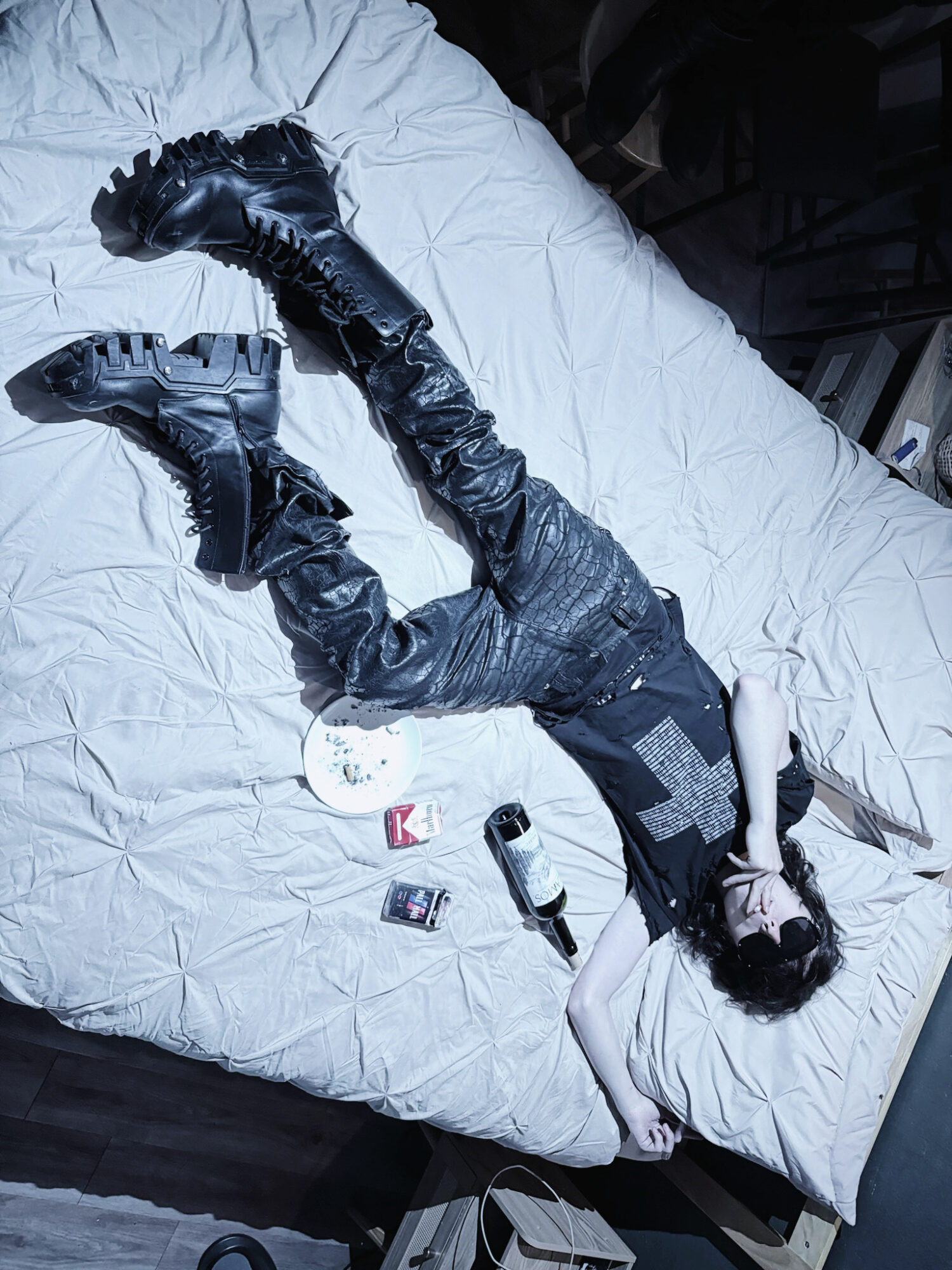 Person lying on bed wearing black clothing and boots, with items scattered nearby, viewed from above.