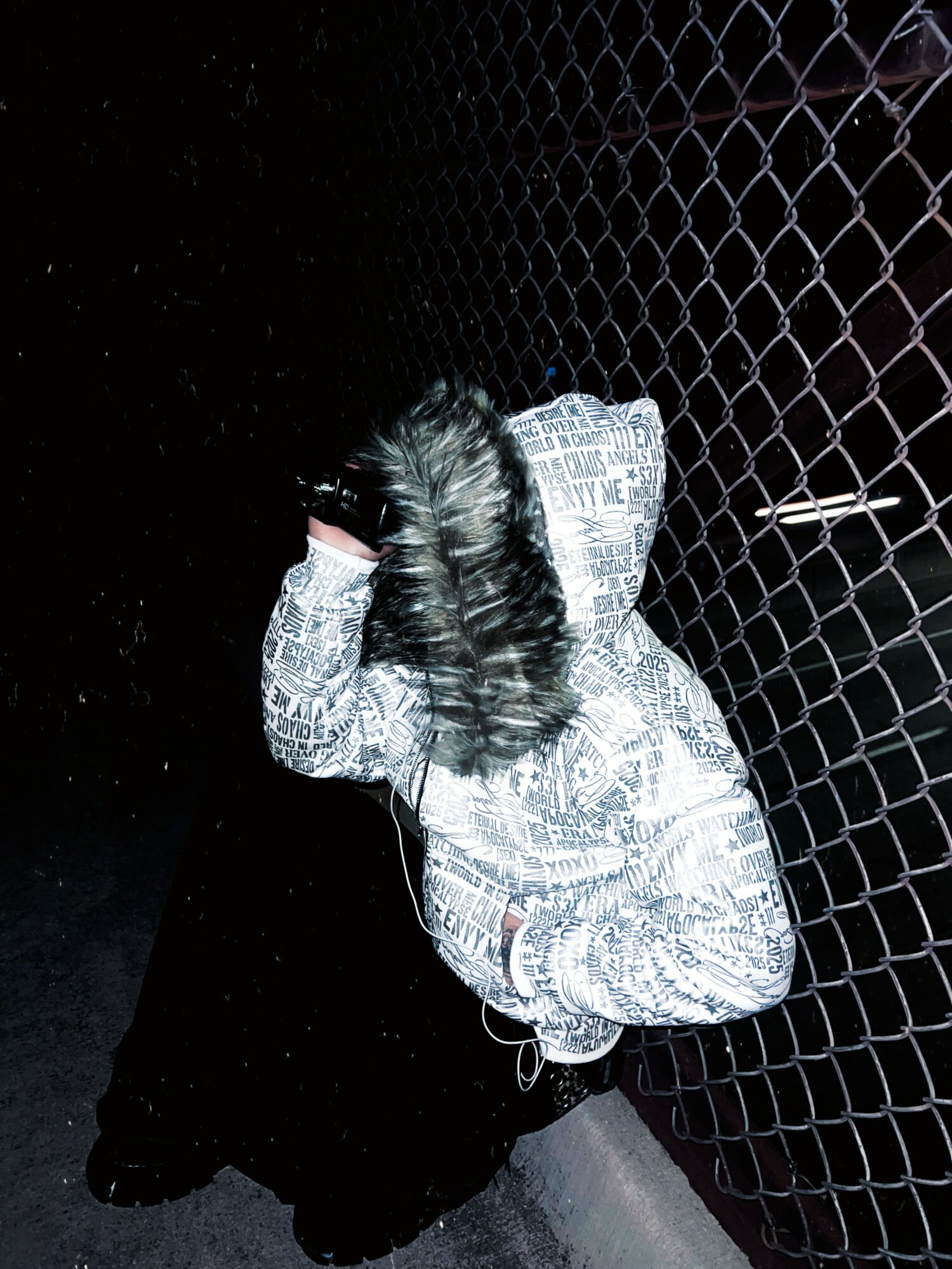 Person crouching with hood up and face hidden, next to a chain-link fence at night.