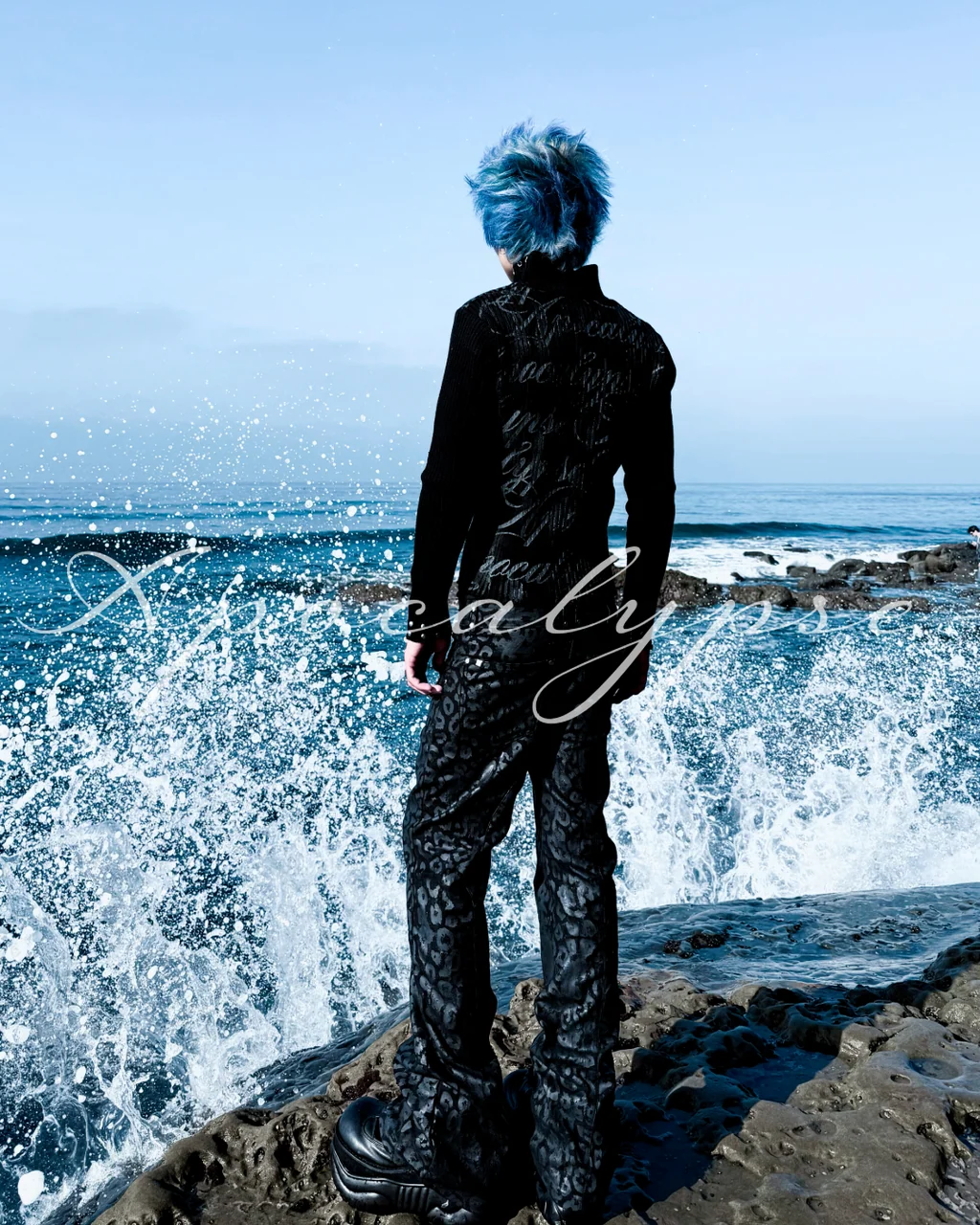 Person with blue hair standing on rocks by the ocean, waves crashing behind them, facing away from the camera.