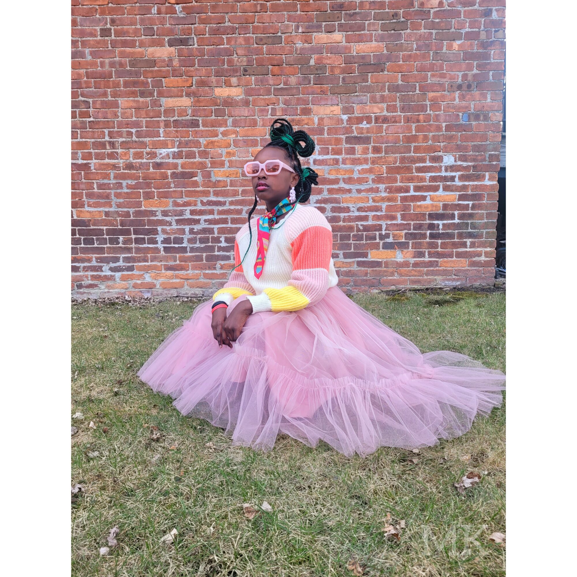 Child sitting on grass in front of a brick wall, wearing pink glasses, colorful sweater, and pink tulle skirt.