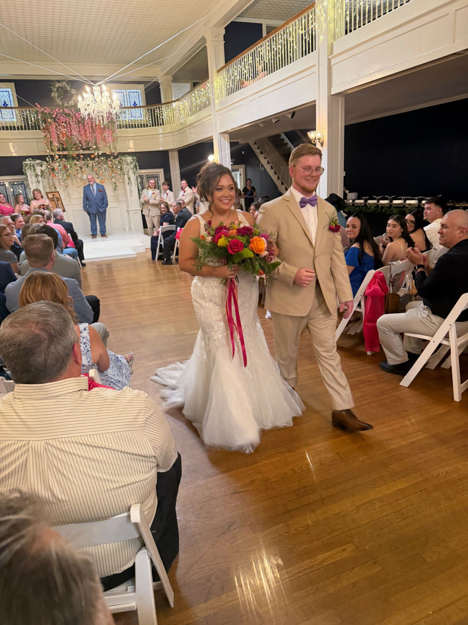 A bride in a white dress and veil holds a bouquet, standing next to a man in a beige suit at a celebration.