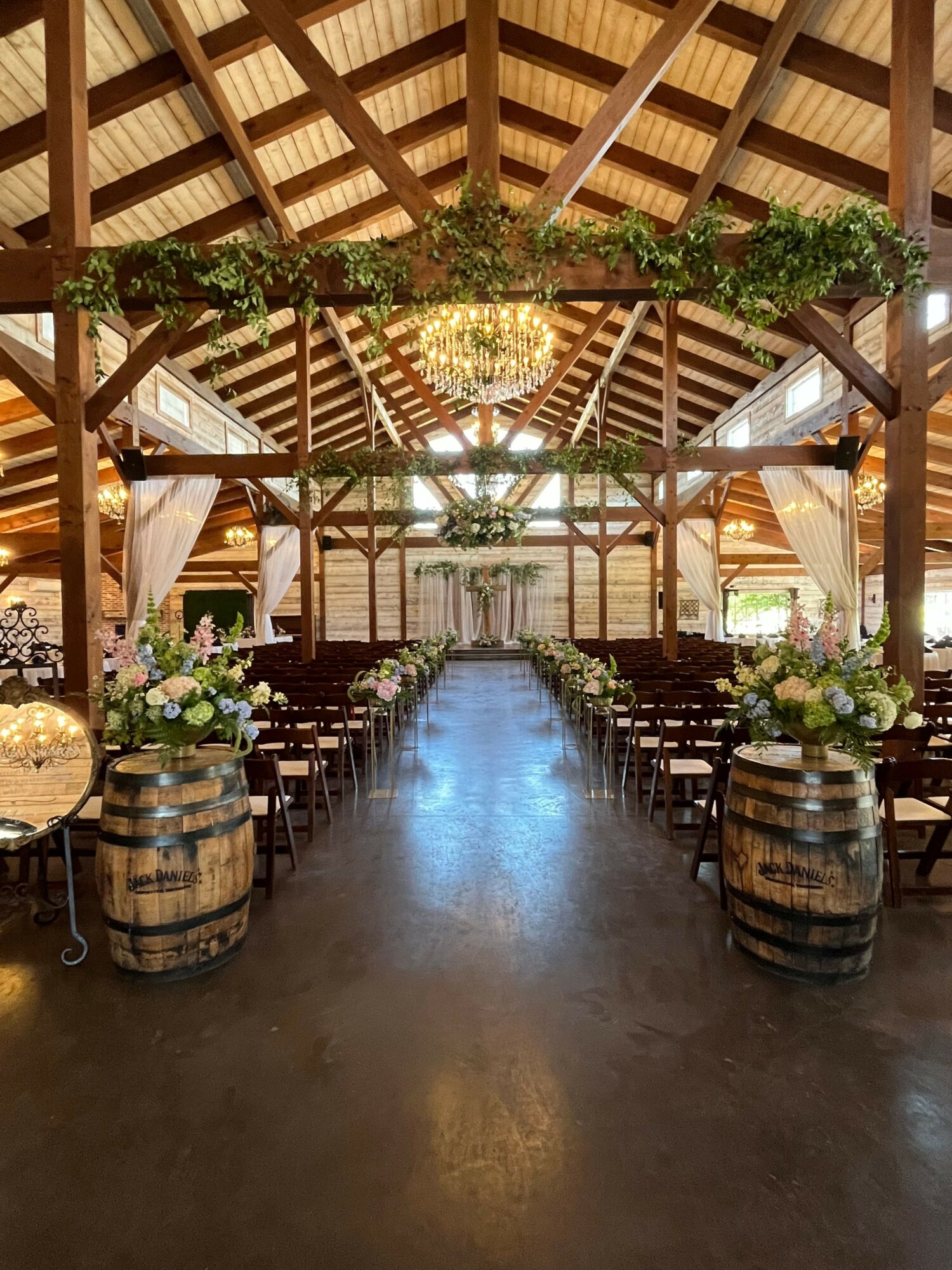 Wooden barn interior decorated with flowers, barrels, and draped fabric, with a central aisle and hanging lights.