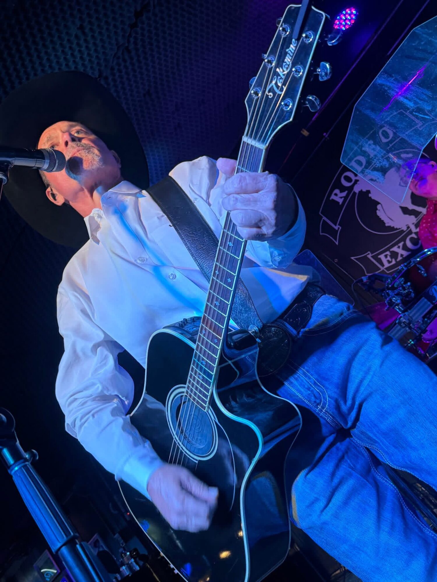 Person wearing a hat playing an acoustic guitar on stage with a microphone, dark background, colorful lights.