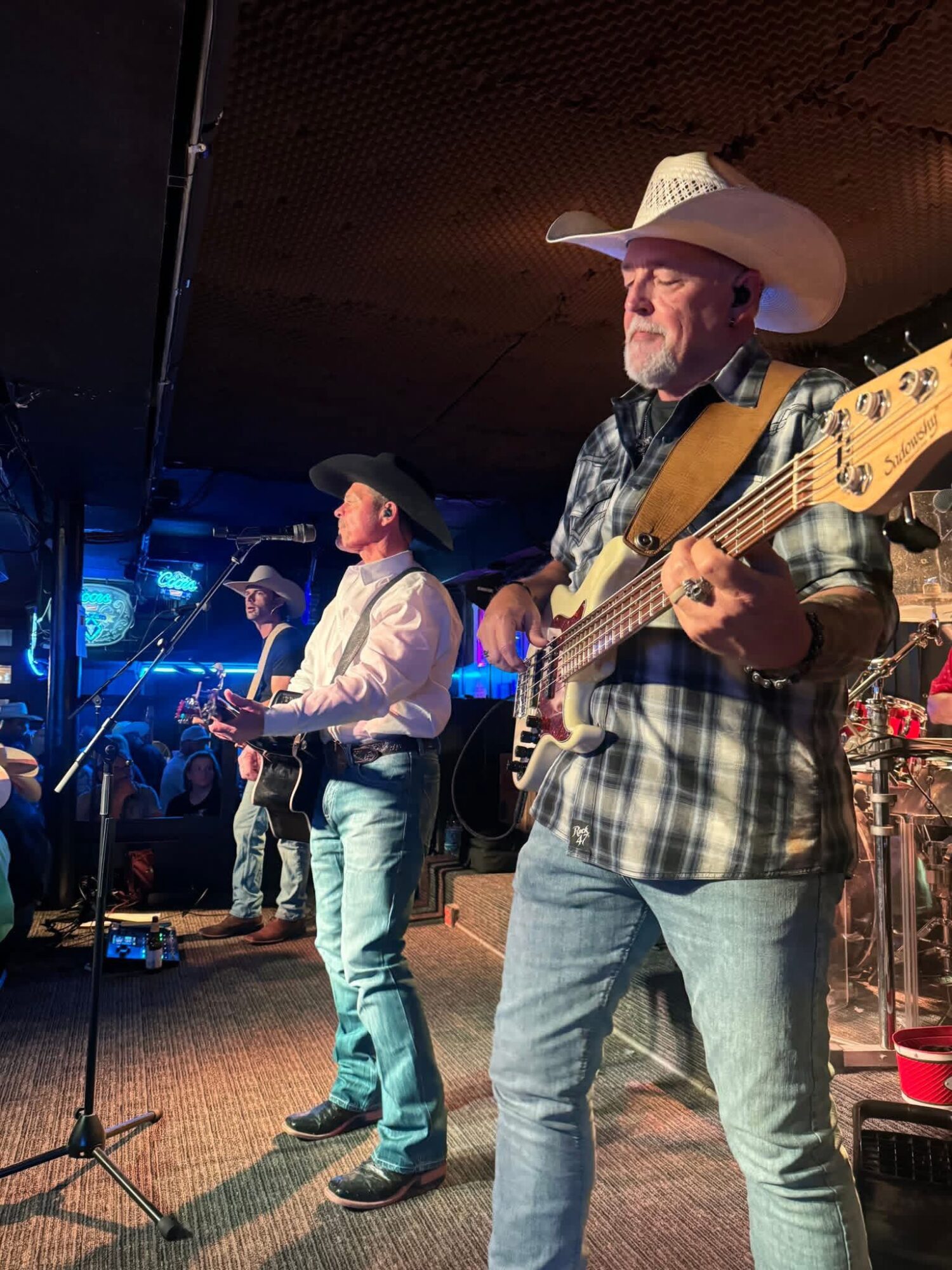 Two musicians performing on stage, one playing guitar and the other singing, both wearing cowboy hats, in a dimly lit venue.