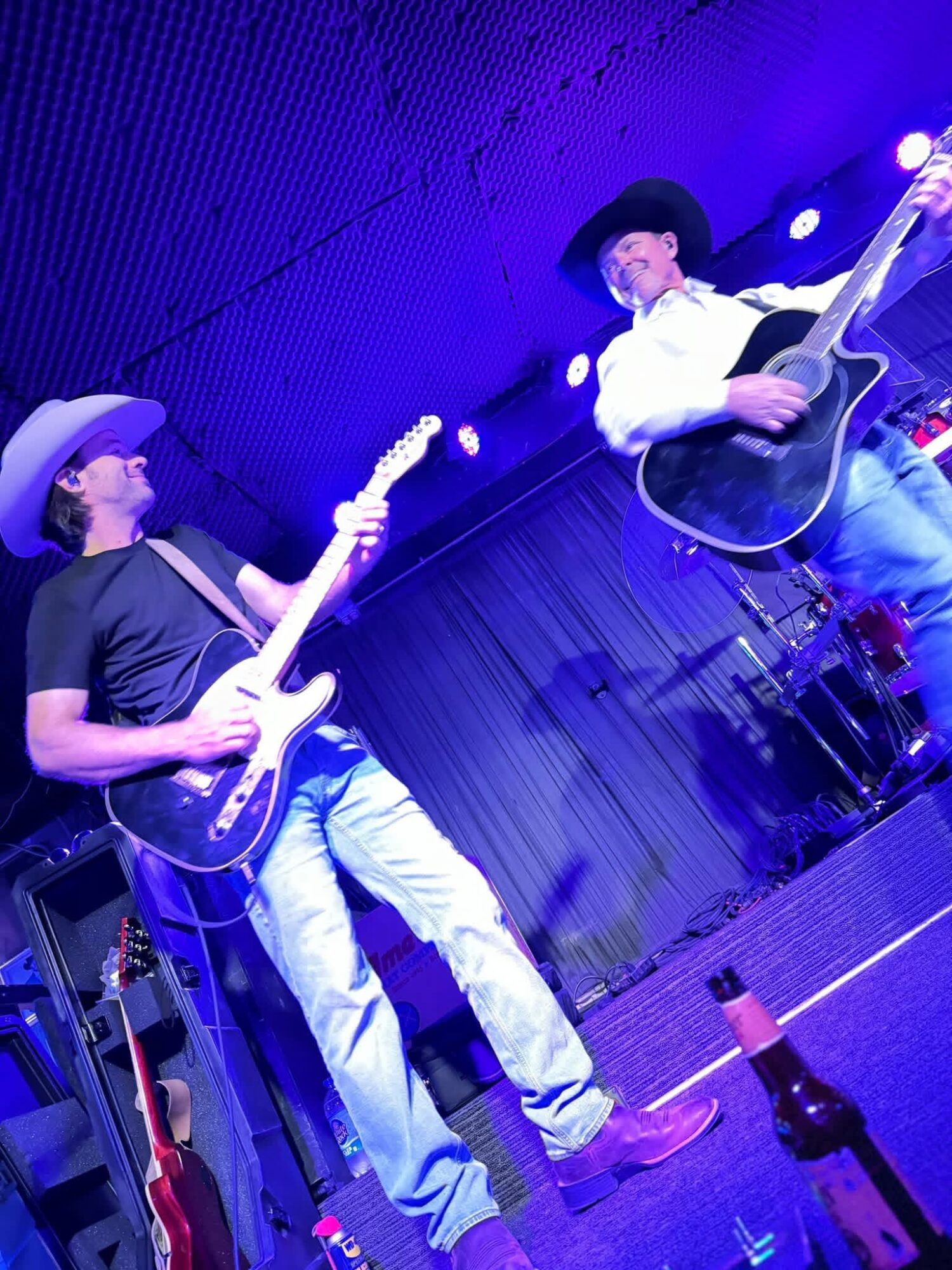 Two musicians playing guitars on stage with blue lighting, one wearing a cowboy hat, the other a black hat, and a bottle on the floor.