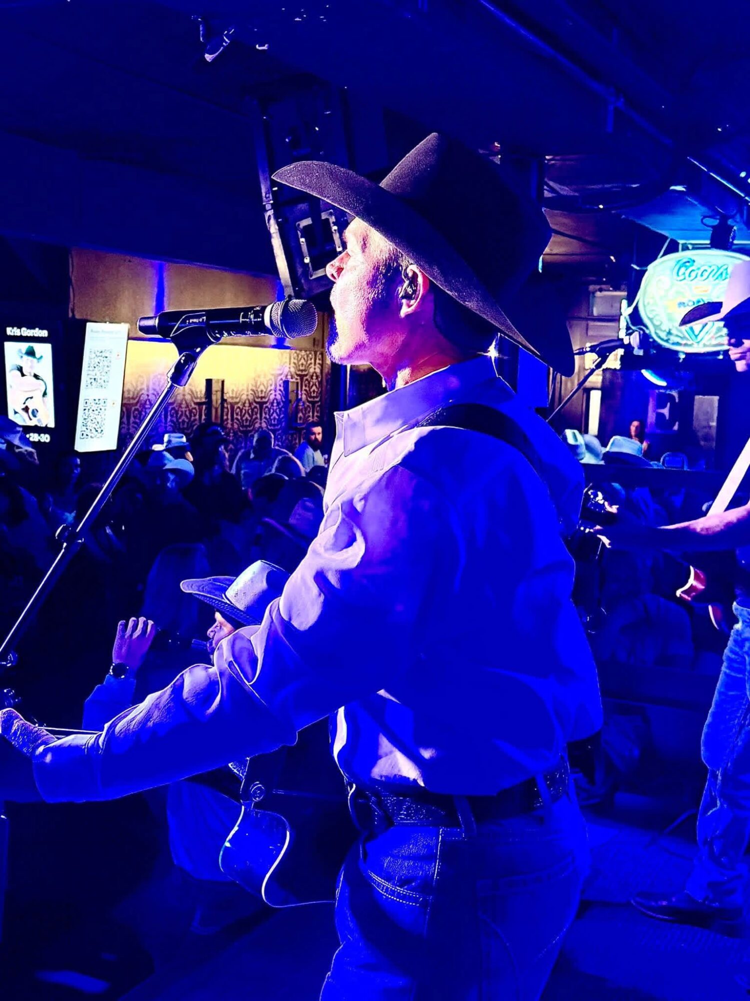 Person wearing a wide-brimmed hat and white shirt performing on stage with microphone, audience in background.