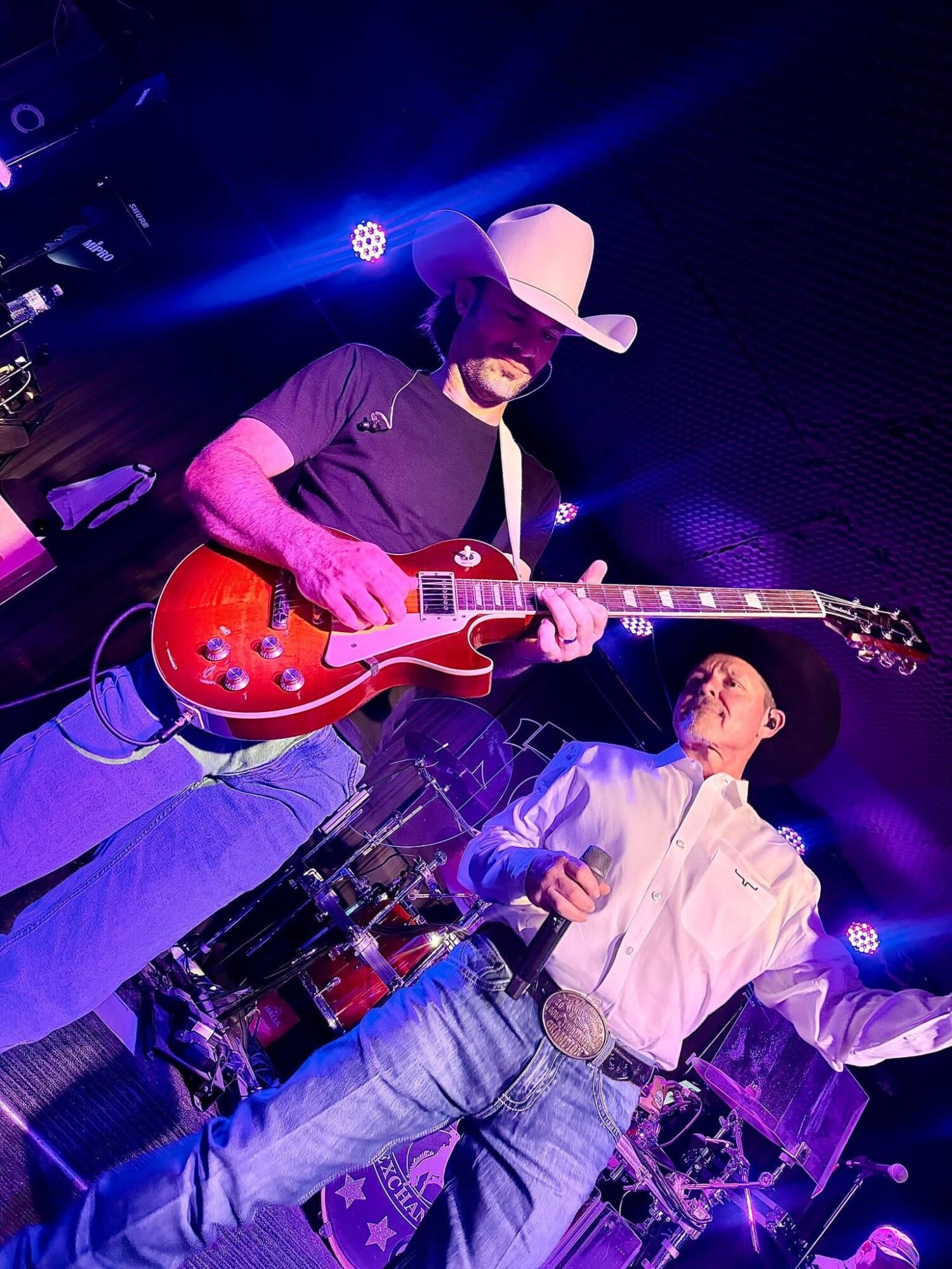 Person wearing a cowboy hat playing a red electric guitar on stage, with another person in a white shirt nearby, stage lights visible.