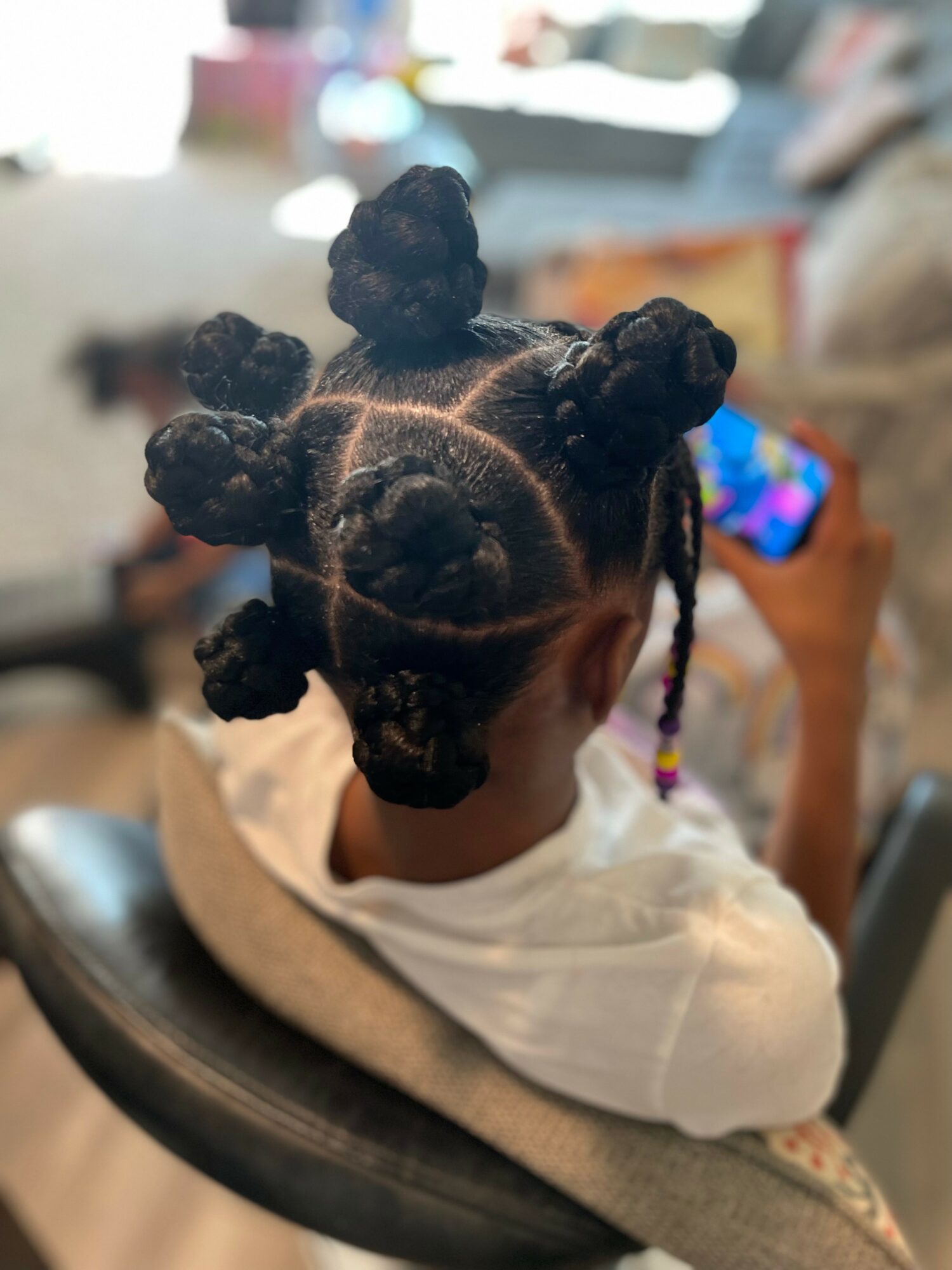 Child with styled hair in multiple bantu knots, sitting in a chair, holding a phone, in a living room.