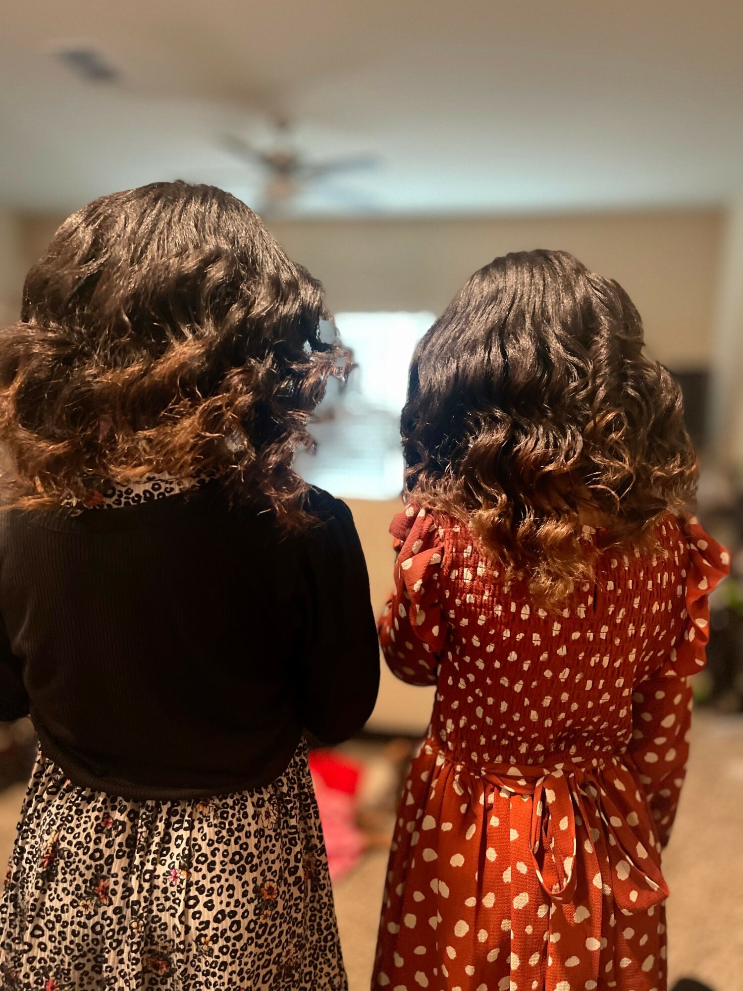 Two people with curly hair standing indoors, facing away from the camera, with a blurred background.