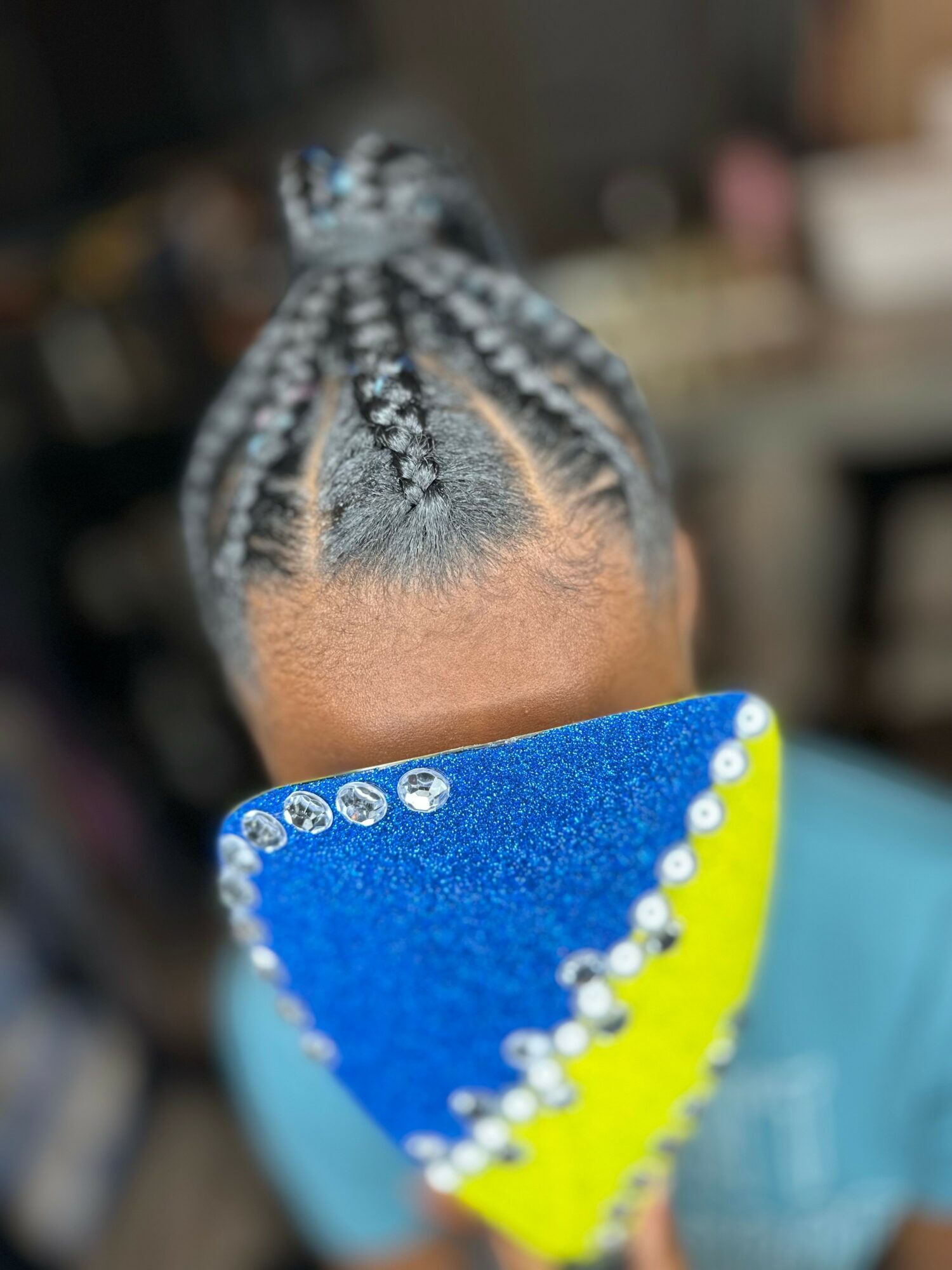 Child with braided hairstyle holding a colorful fan, face partially covered, indoors with blurred background.