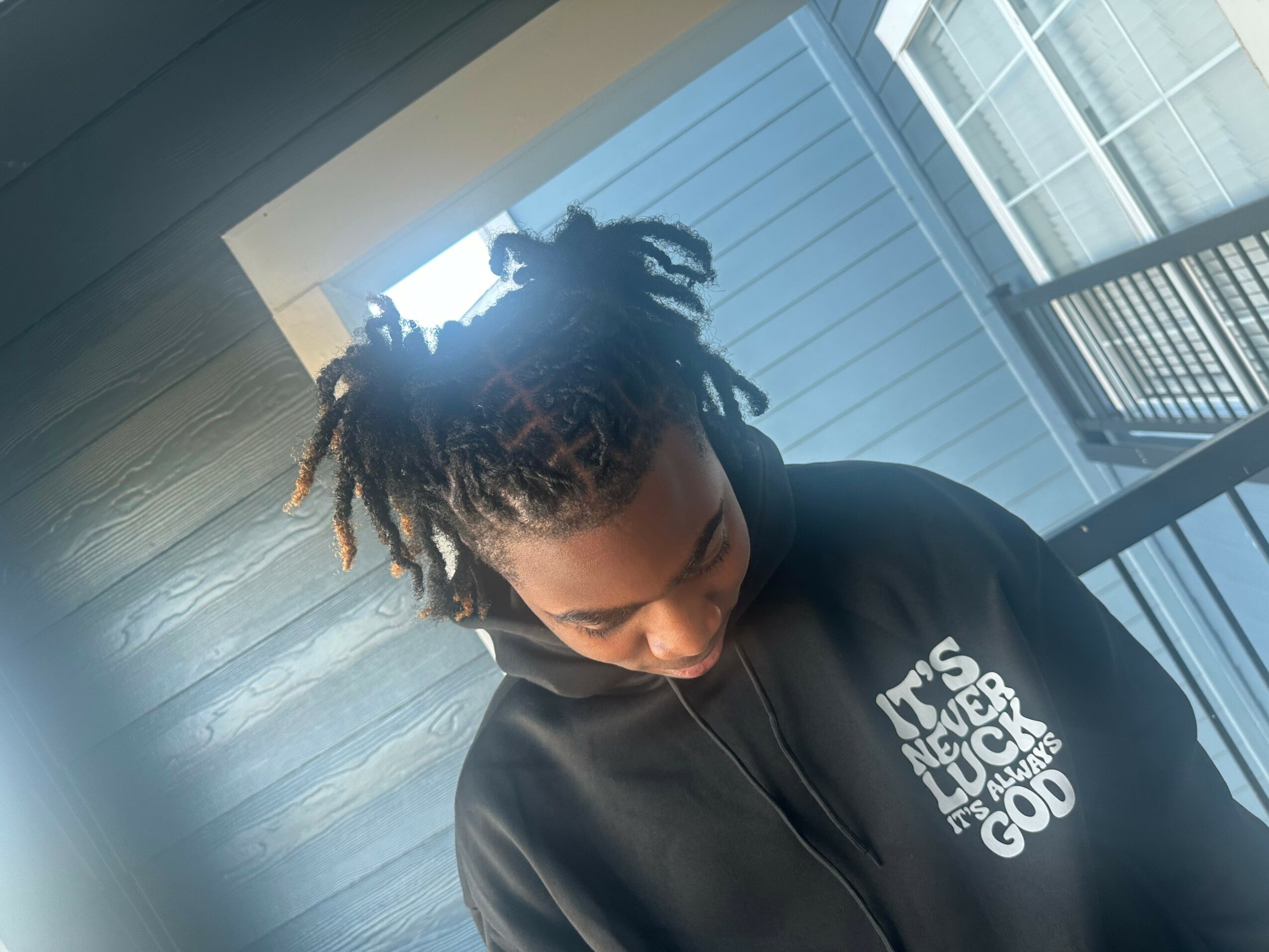 Young person with dreadlocks looking down, wearing a black hoodie with white text, standing indoors near a window.