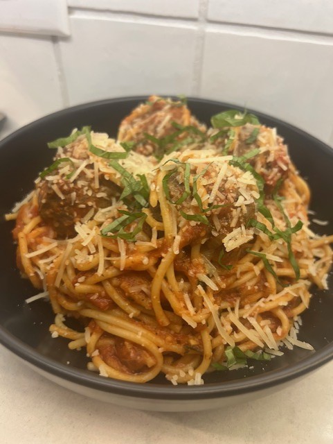 Plate of spaghetti with meatballs, topped with grated cheese and chopped herbs, on a black dish against a white tiled background.