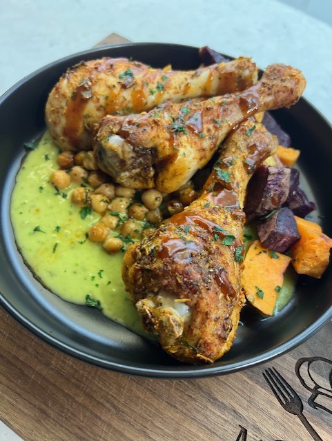 Plate with roasted chicken drumsticks, chickpeas, and roasted vegetables on a black dish, served on a wooden table.