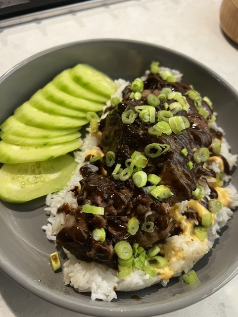 Dish with sliced avocado and rice topped with dark sauce, chopped green onions, and nuts on a gray plate.