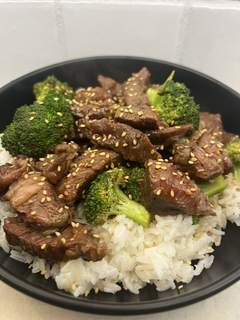 Beef stir-fry with broccoli and sesame seeds served over white rice in a black bowl.